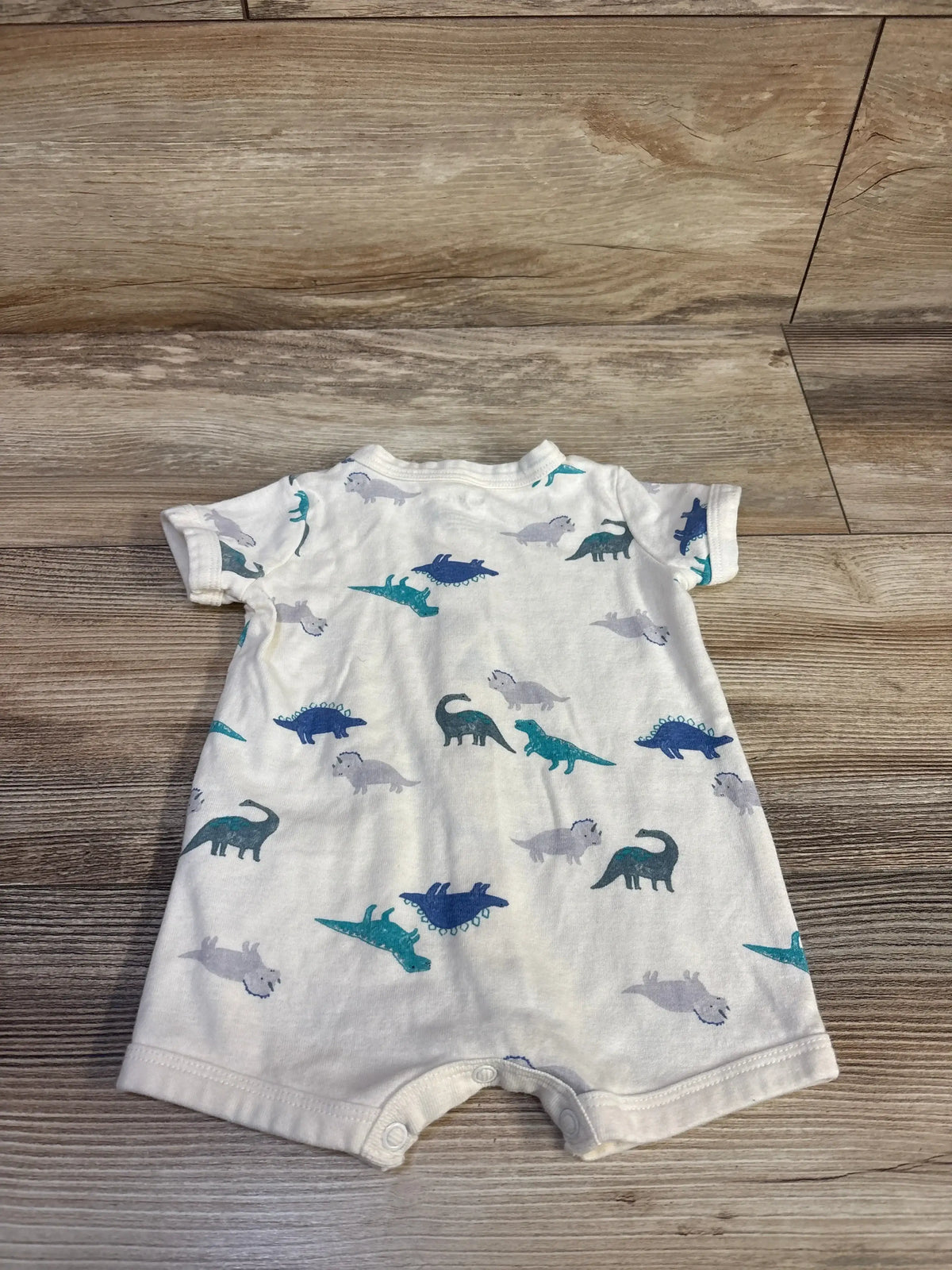 Front view of Carter’s white Dinosaur Shortie Romper showcasing dinosaur print, short sleeves, and snap crotch on wood floor