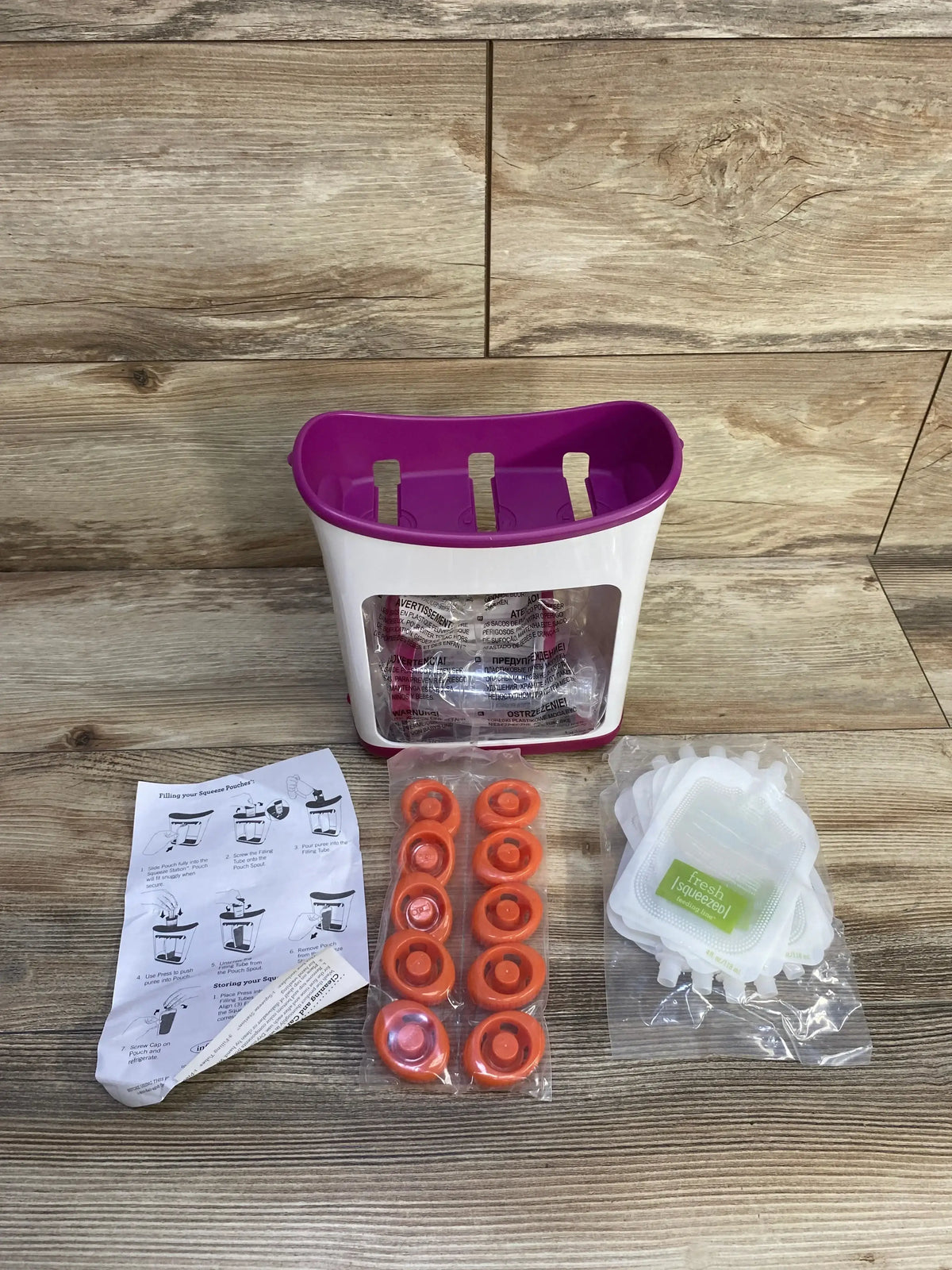 Front view of Infantino Squeeze Station with purple top, clear front, orange sealing rings, pouches and instructions on wood