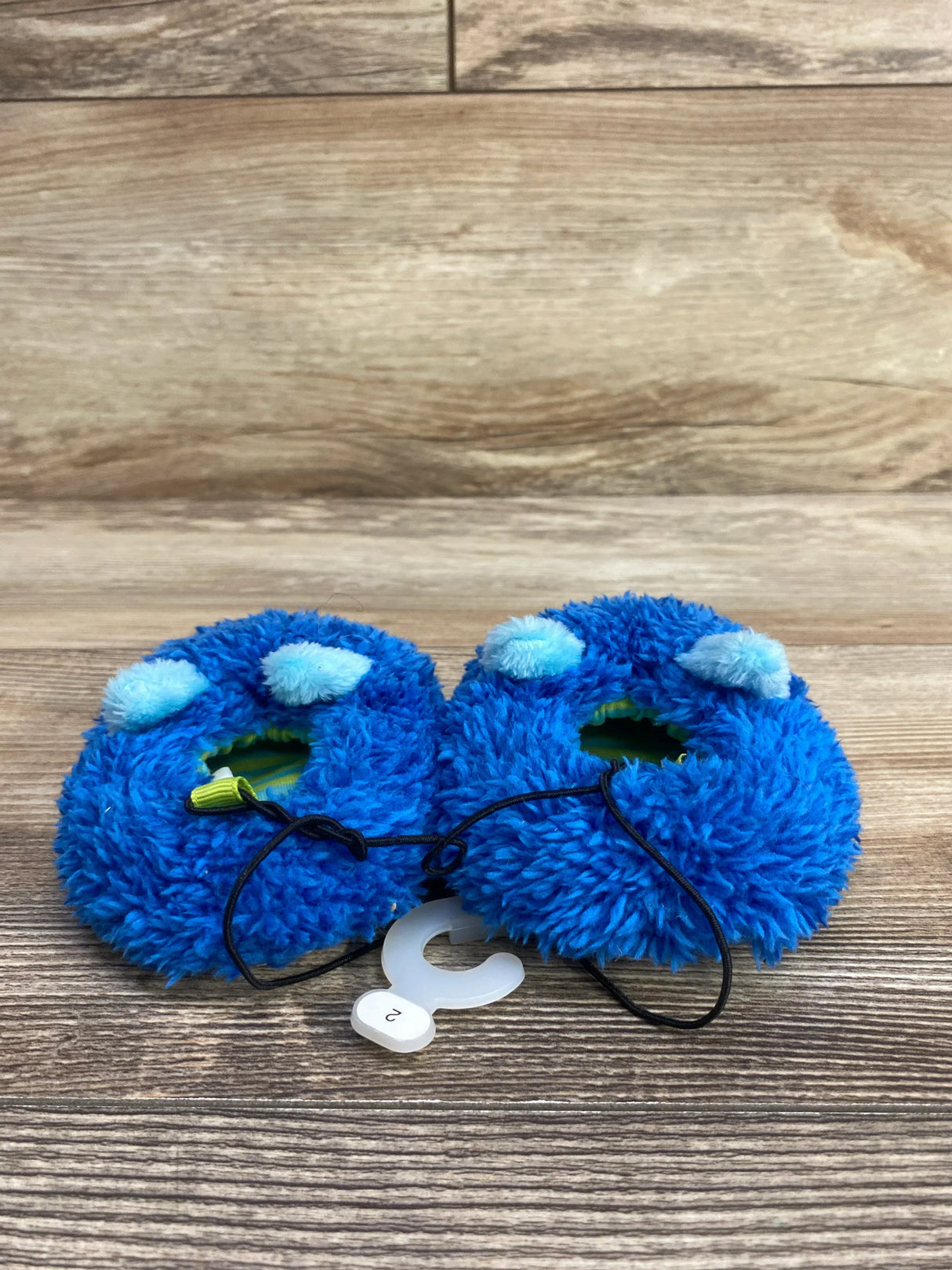 Pair of blue fuzzy baby booties with pom-poms on wooden surface, size 2 tag attached