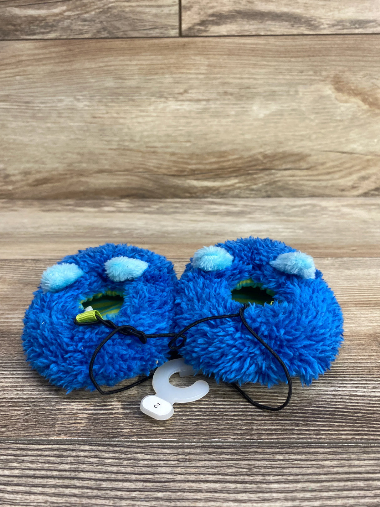 Pair of blue fuzzy baby booties with pom-poms on wooden surface, size 2 tag attached
