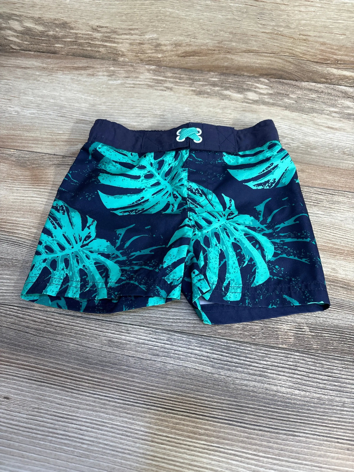 Top-down view of Cat and Jack navy swim trunks 12M with teal tropical leaf print and drawstring waist on wood background