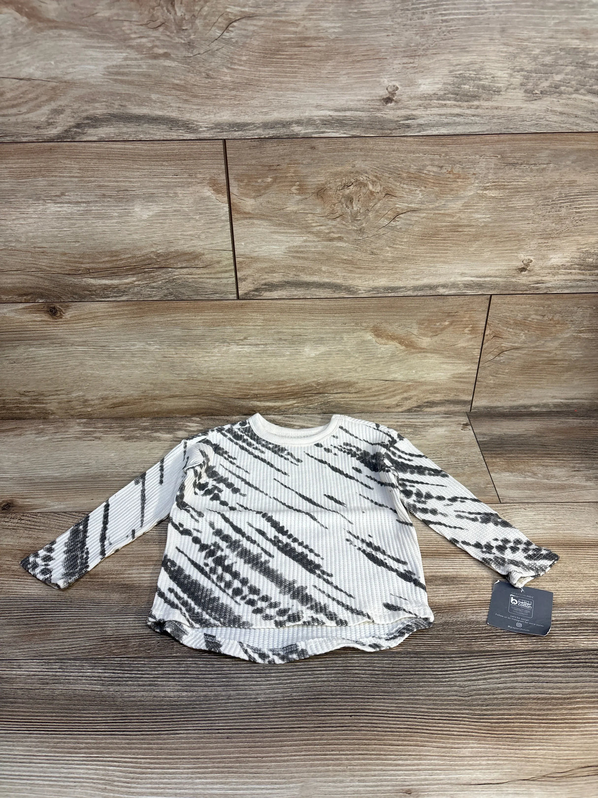 Flat lay of NEW Grayson Mini Waffle Knit Shirt in ivory showcasing black tie-dye pattern with hangtag on wood plank floor