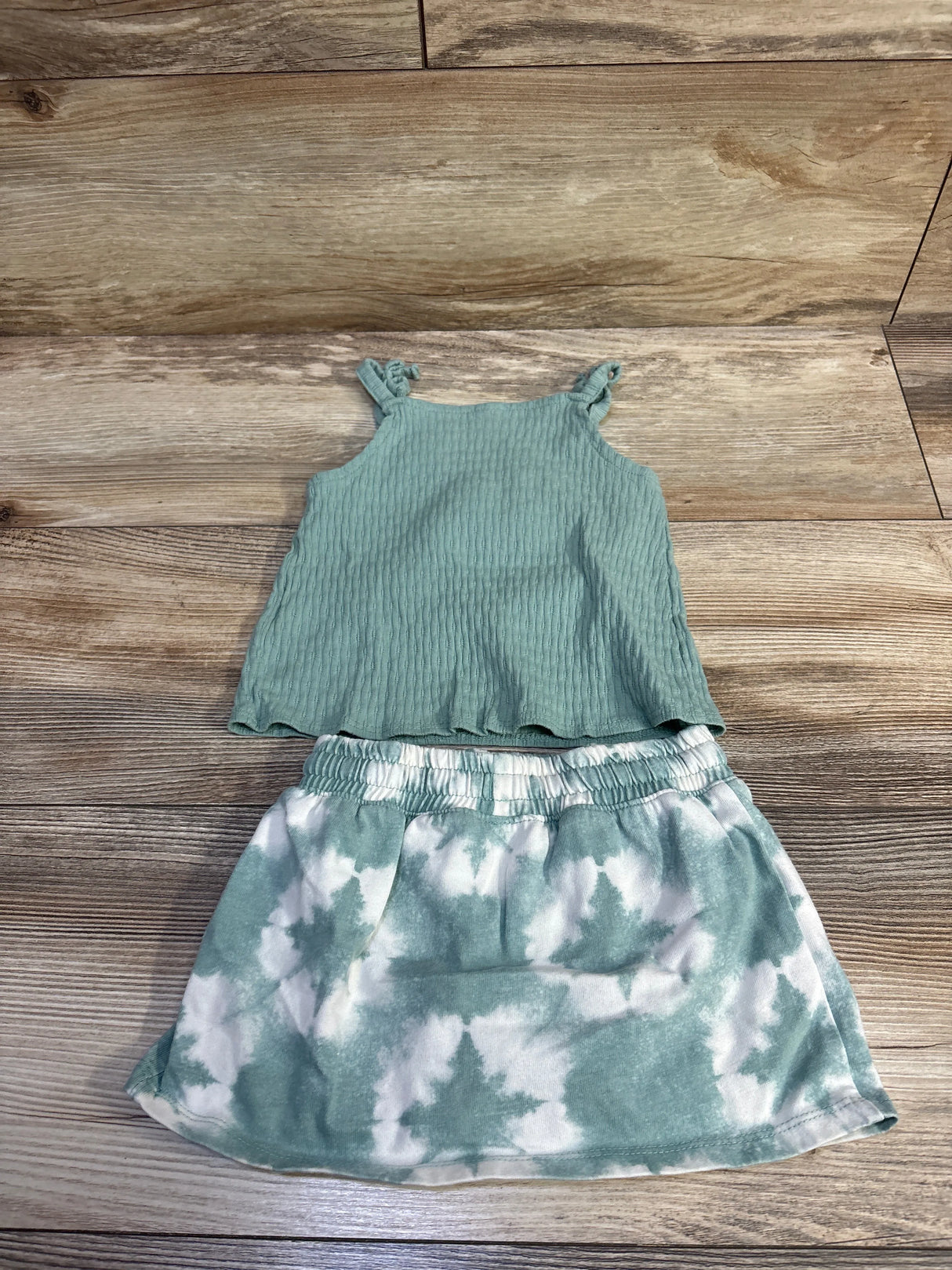 Top down view of toddler green ribbed tank with knotted straps and matching green and white tie dye skort with elastic waistband on wood background