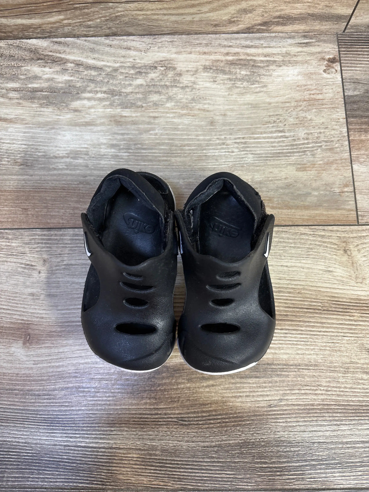 Top-down view of Nike Sunray Protect 3 toddler black sandals showing three toe vents, Velcro heel straps, and white sole edges on wood floor