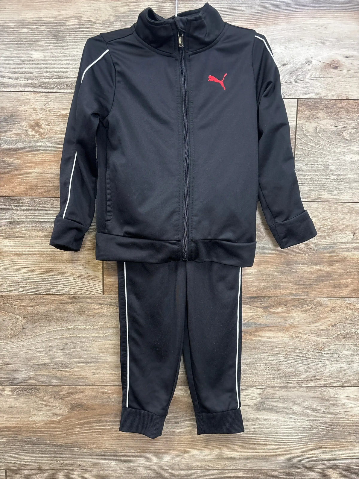 Black kids athletic tracksuit with red logo and white stripes, displayed on wood background