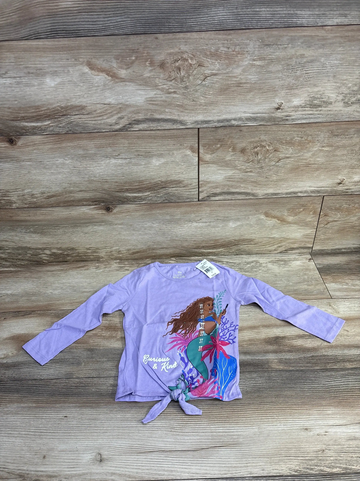 Lilac long-sleeve toddler shirt with mermaid graphic and 'Curious & Kind' text on wooden floor