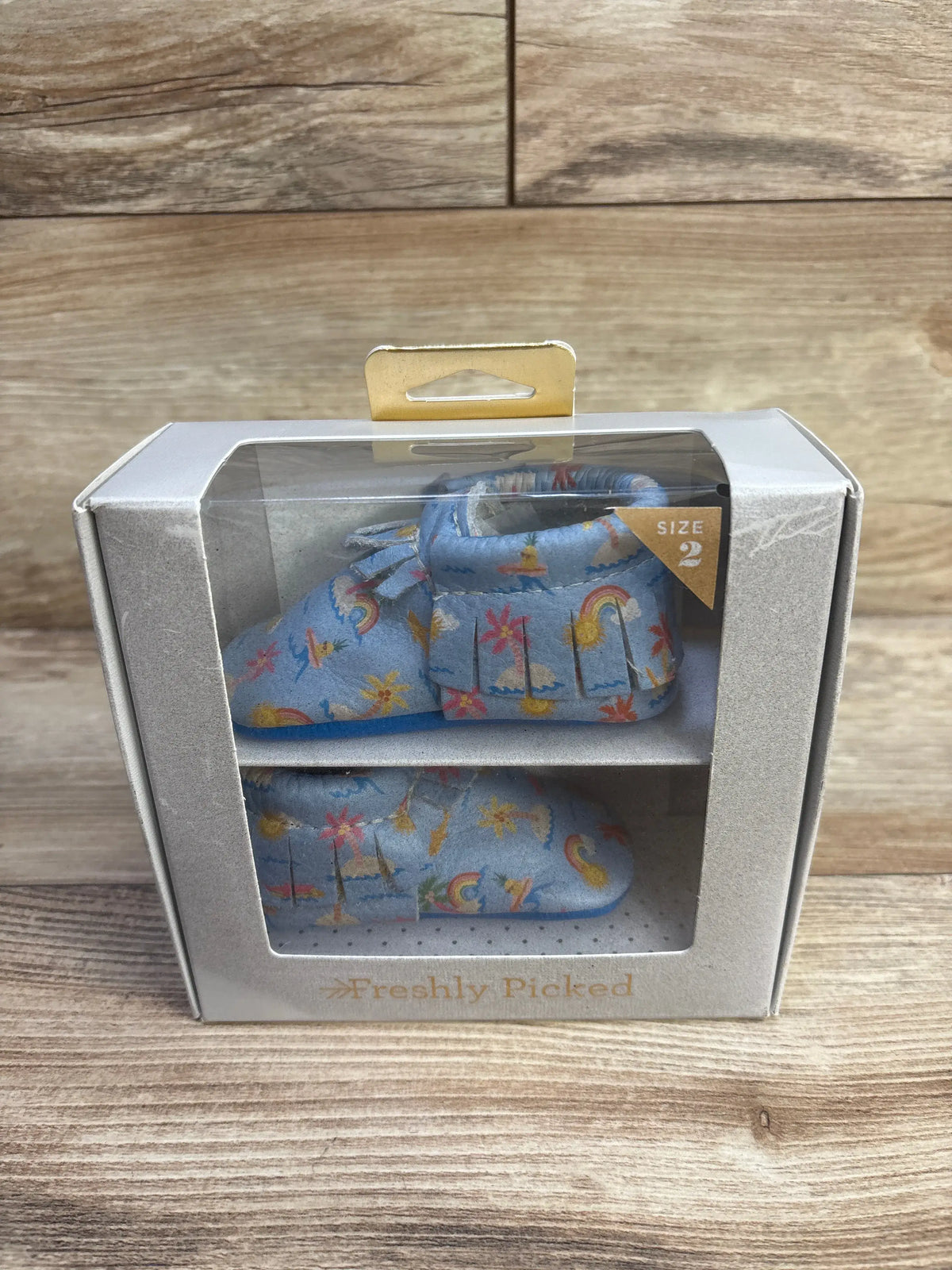 Front and side view of Freshly Picked blue tropical print baby moccasin showing fringe trim and size 2 tag in box on wood background