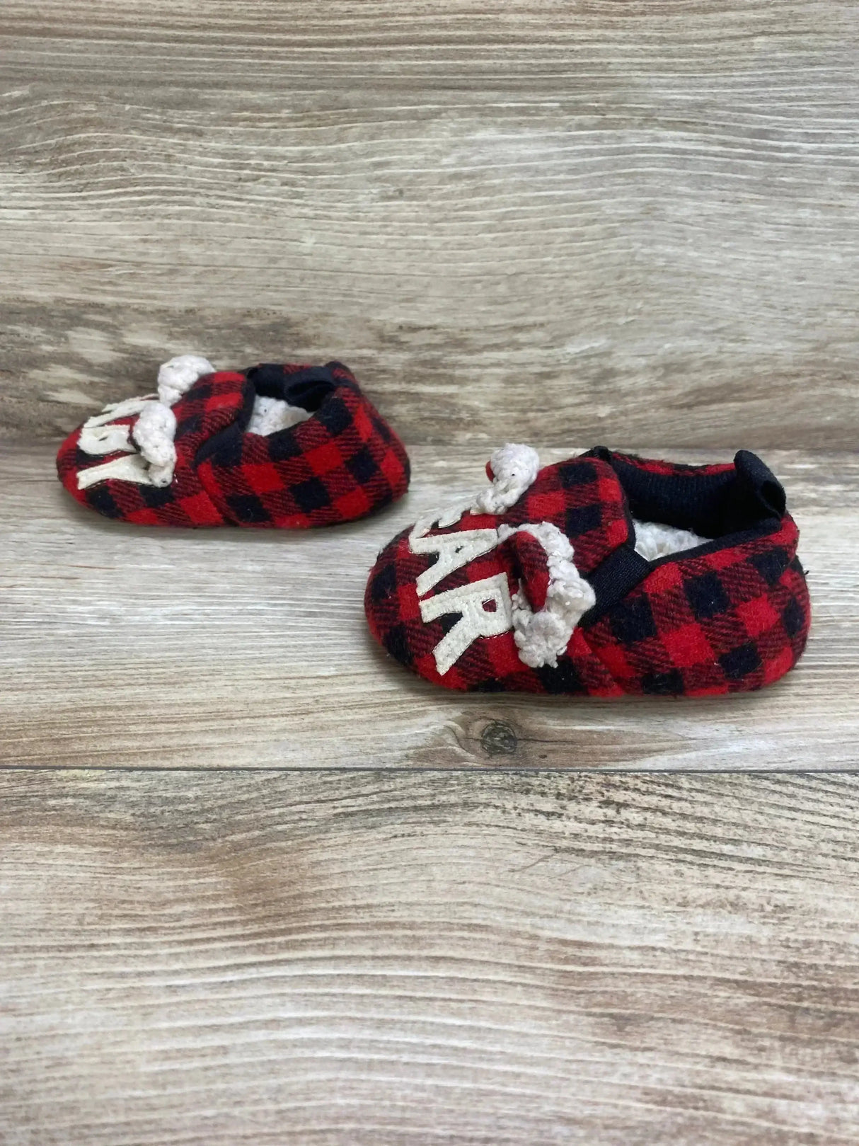 Front-angled view of Buffalo baby bear slippers size 1C showcasing red and black buffalo plaid, white bear face detail and fleece lining on wood-look background