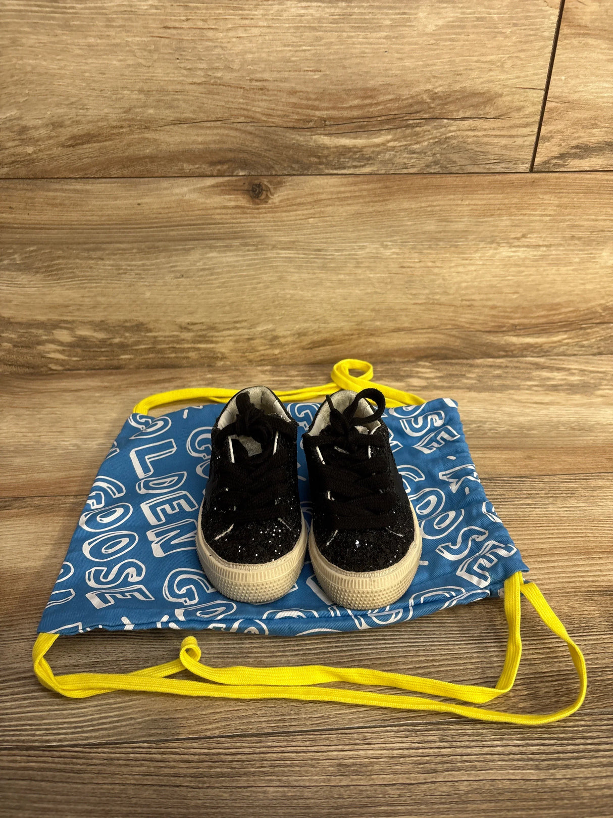 Pair of black glitter kids sneakers on blue drawstring bag with white text and yellow strings on wooden surface