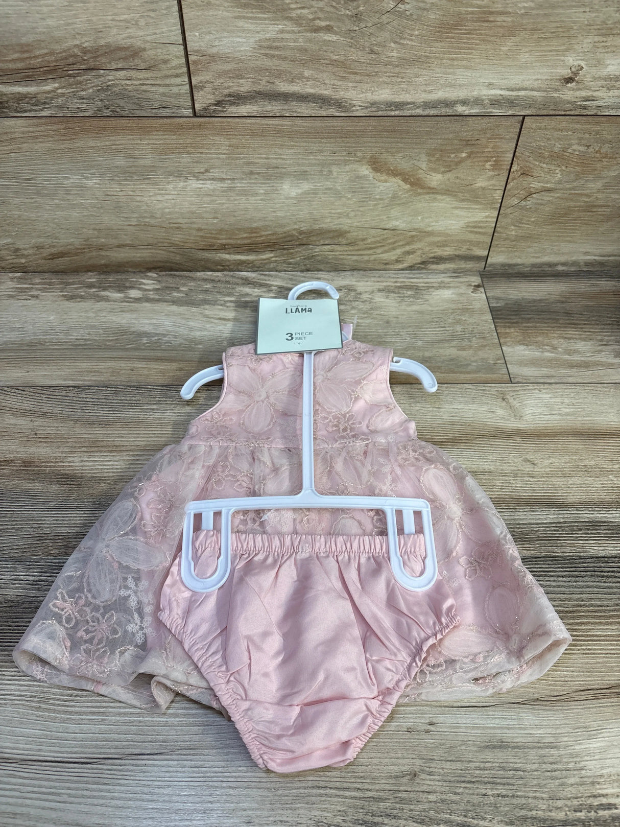 Flat-lay of pink sleeveless baby dress with floral lace overlay and matching satin bloomers on wood floor, size 3M