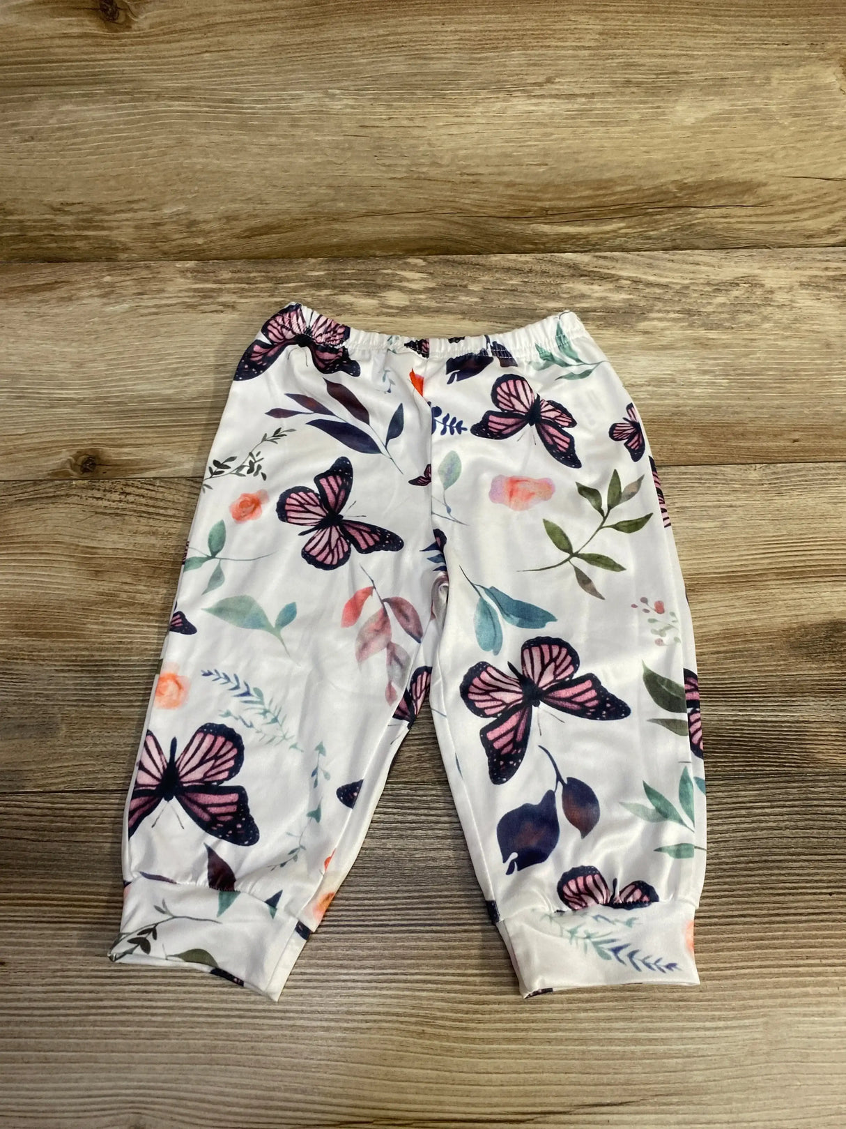 Flat lay of white baby pants (6–9 months) showcasing pink and navy butterfly print, elastic waistband and cuffed ankles on wood background