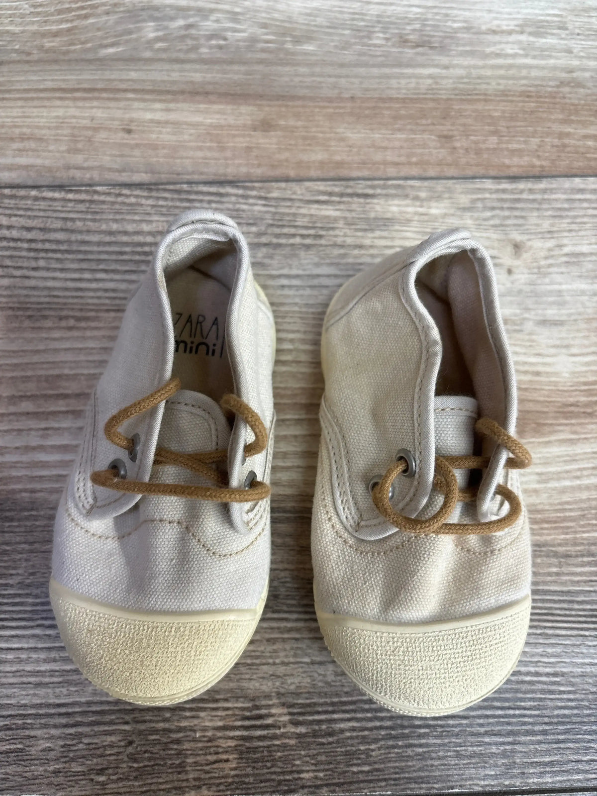 Pair of beige toddler canvas shoes with brown laces on wooden surface, kids' casual footwear