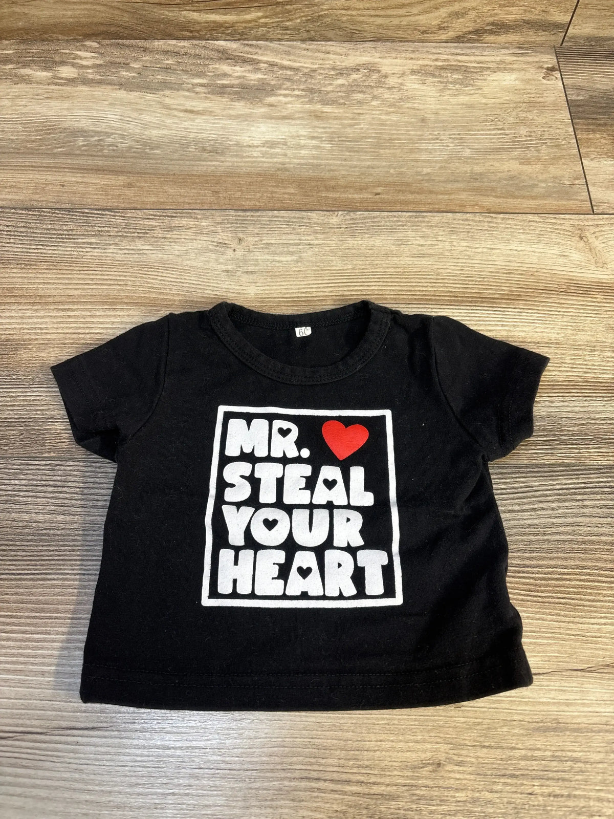 Black baby t-shirt with white text 'Mr. Steal Your Heart' and red heart graphic on wood surface