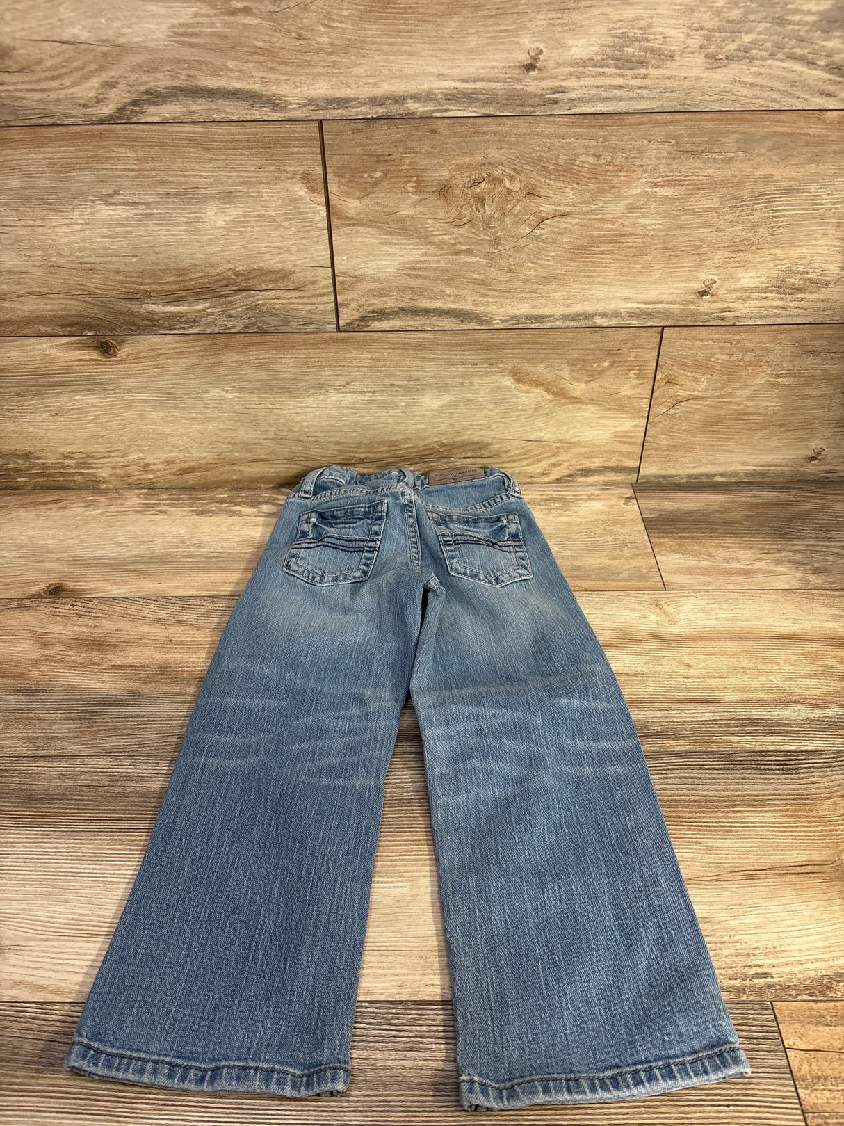 Flat lay of boys' light wash relaxed-boot denim jeans showing rear pocket stitching and faded whiskering on wood floor