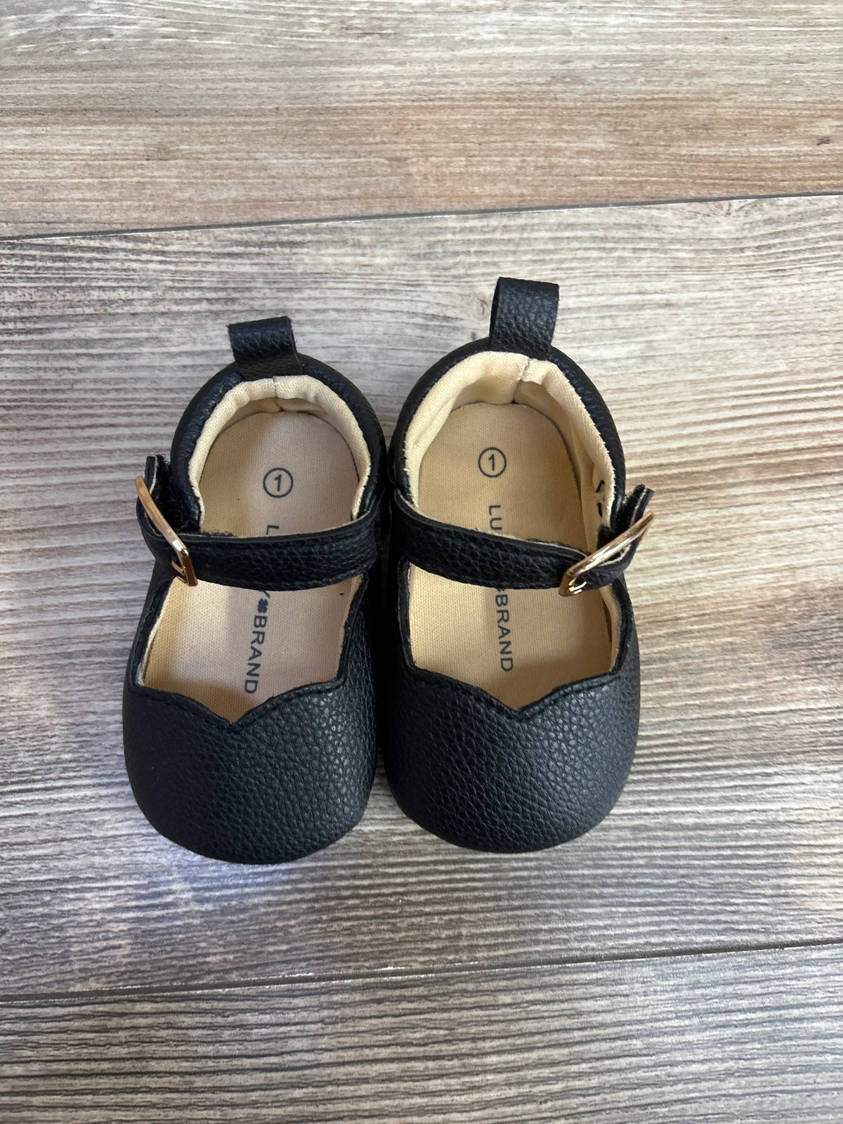 Top view of Lucky Brand Mary Jane crib shoes in black, with ankle straps and gold buckles on wood grain