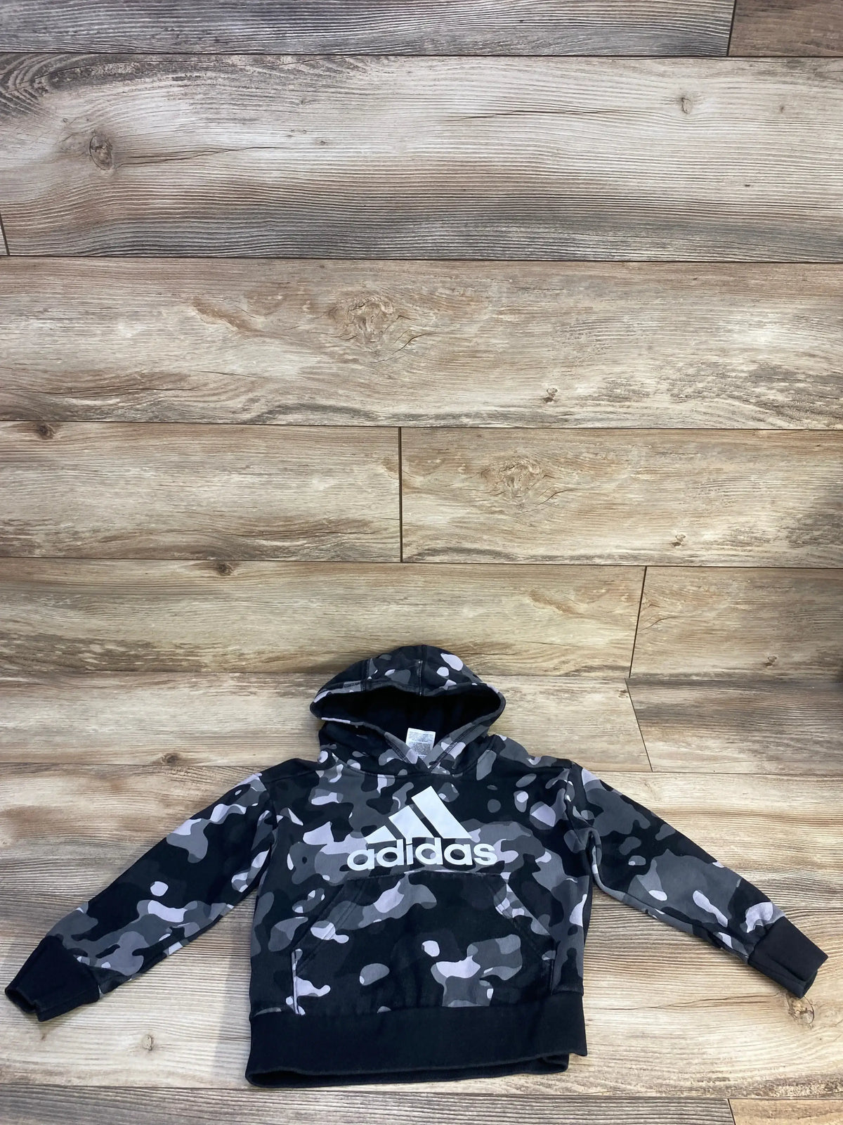 Front view of toddler Adidas pullover sweatshirt in grey camo showcasing white adidas logo, hood, and kangaroo pocket on wood floor background