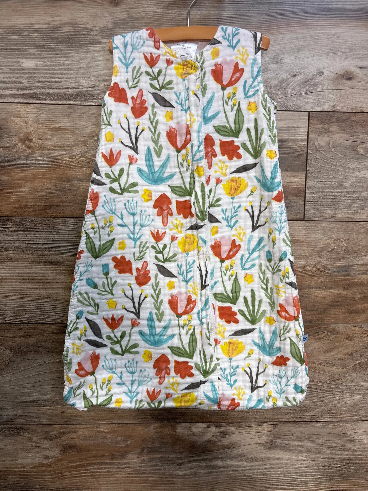 Front view of Little Unicorn cotton muslin sleep sack showcasing a colorful floral print and central zipper on a wood floor