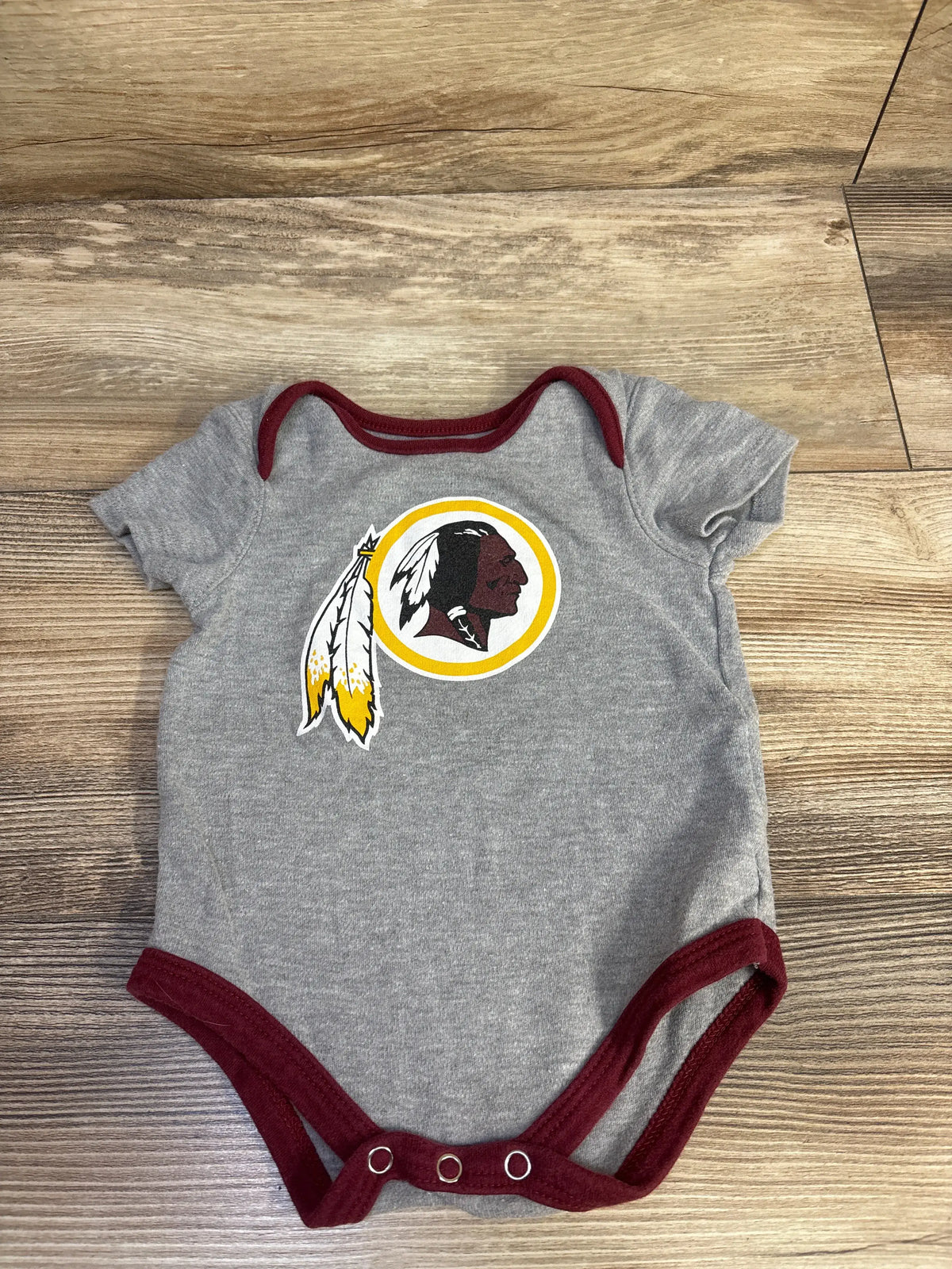 Front view of NFL Washington Redskins grey bodysuit, size 3/6 months, showcasing team logo and snap closure on wood floor