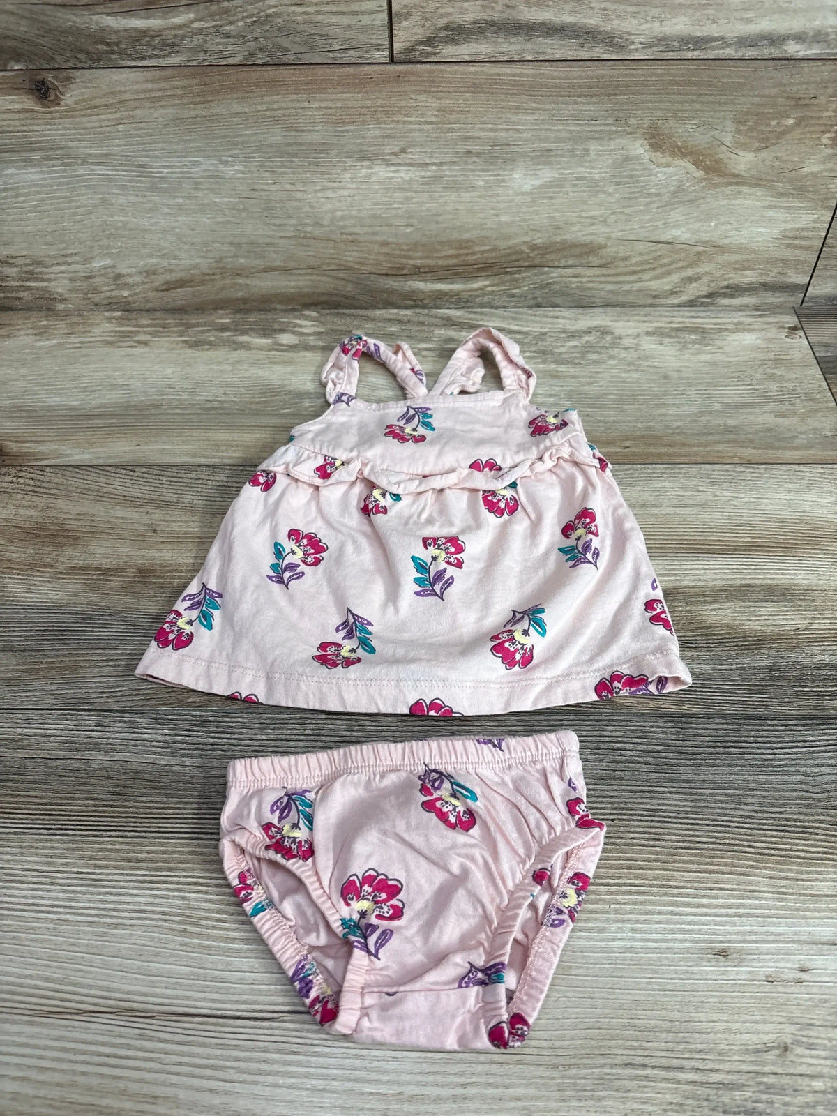 Flat lay of pink Carter’s 2-piece floral dress and bloomers with ruffle detail and magenta-teal flowers on wood