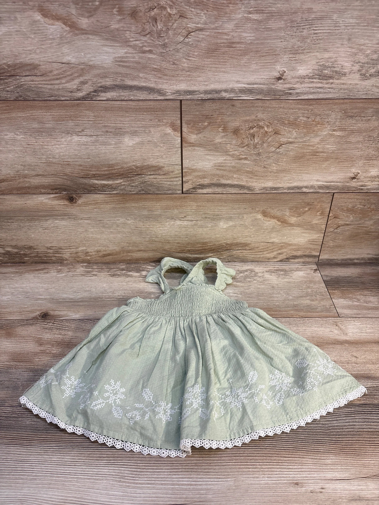 Light green toddler sundress with white floral embroidery and lace trim, laid on wood