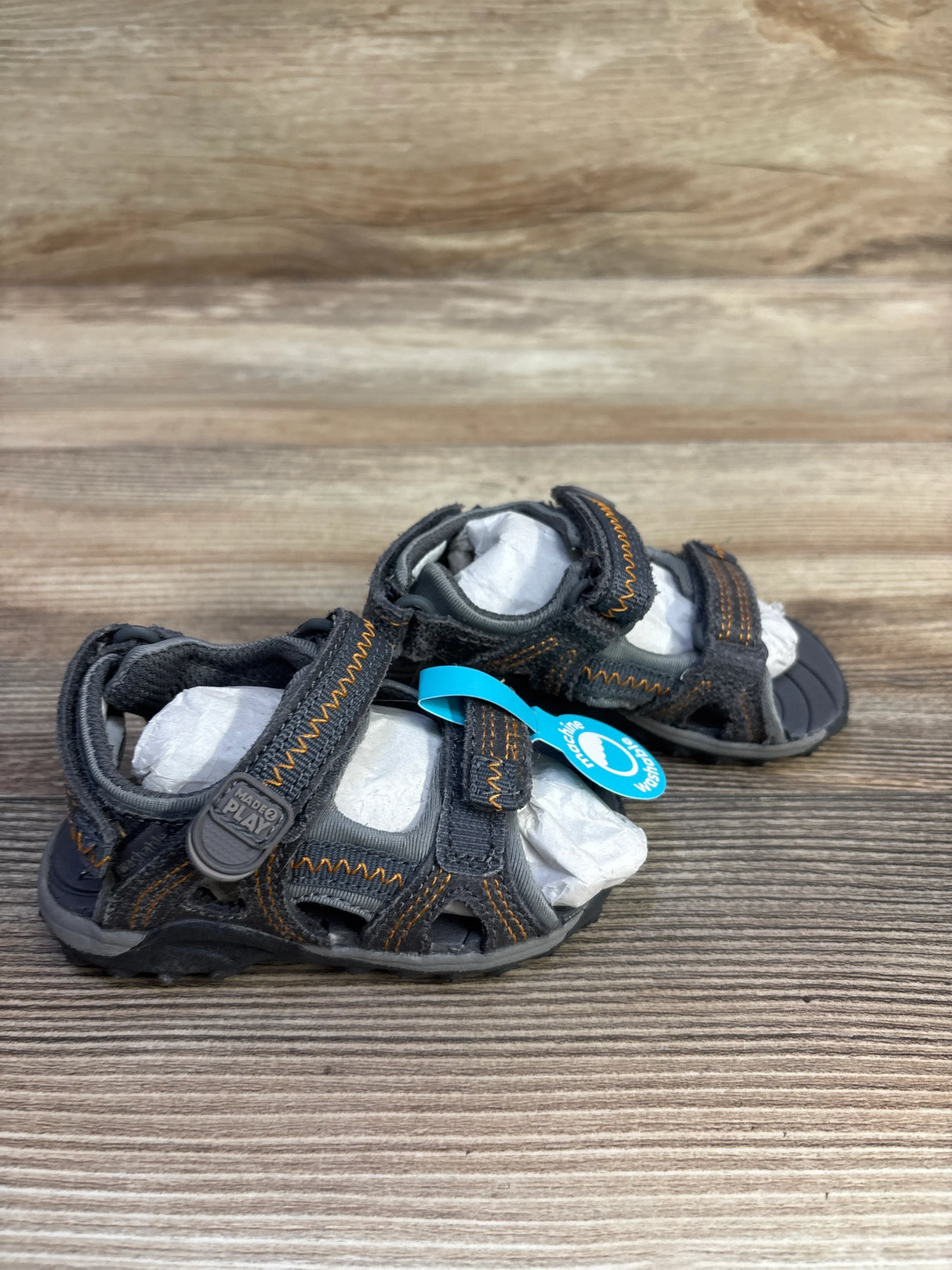 Three-quarter view of Stride Rite Made 2 Play Isaac toddler sandals, grey 5C, with Velcro straps and orange stitching on wood