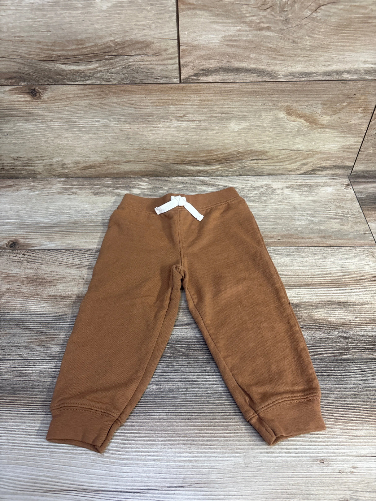 Flat lay of brown toddler drawstring joggers showcasing white drawstring and cuffed ankles on light wood plank background