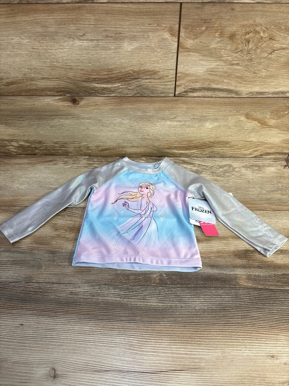 Front flat lay of toddler Elsa long-sleeve rashguard with pastel blue-pink body, Elsa illustration, and iridescent silver sleeves on wooden background