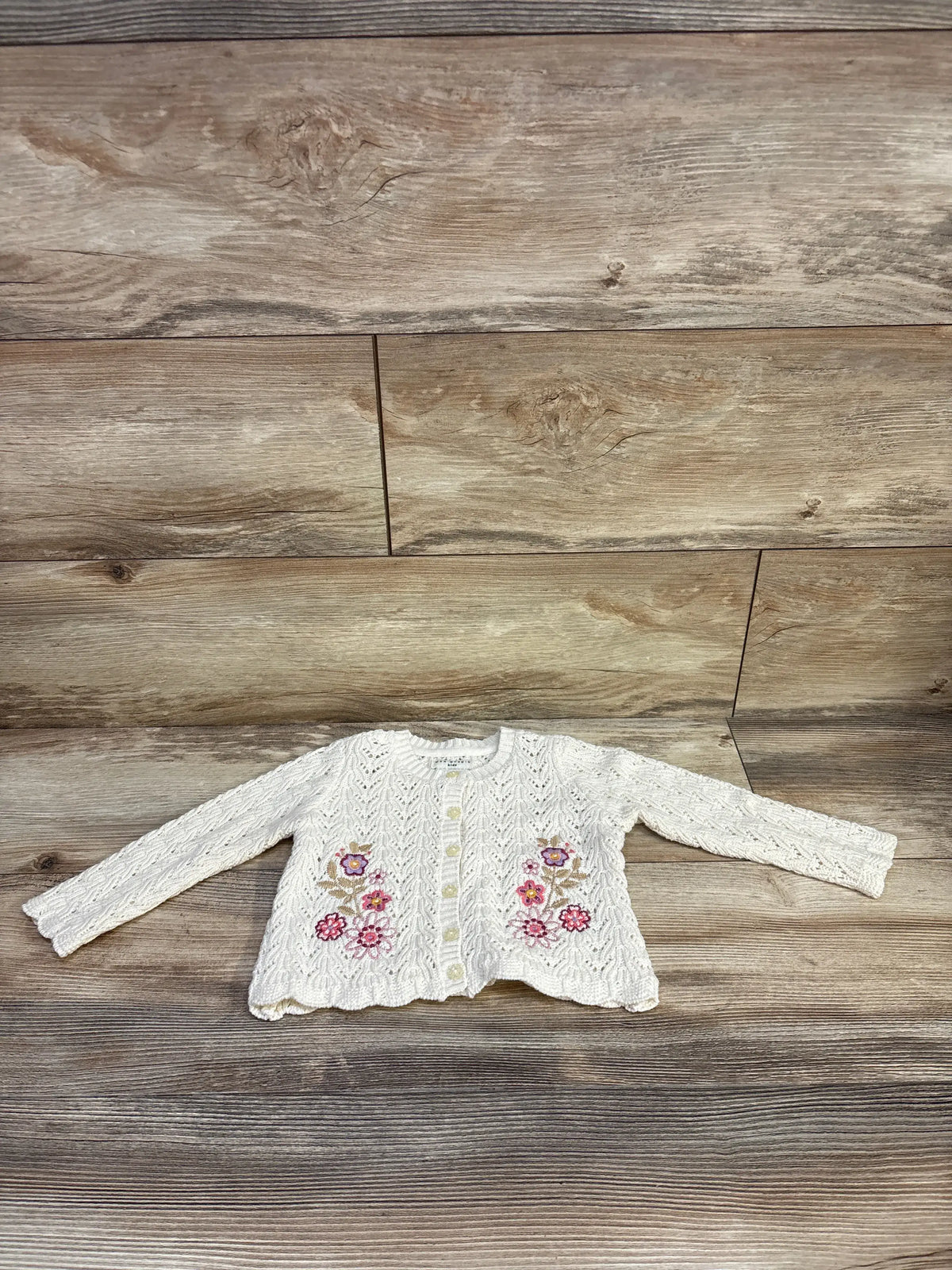 Front view of ivory baby knit cardigan showing embroidered pink and purple flowers, button front, and scalloped hem on wood surface