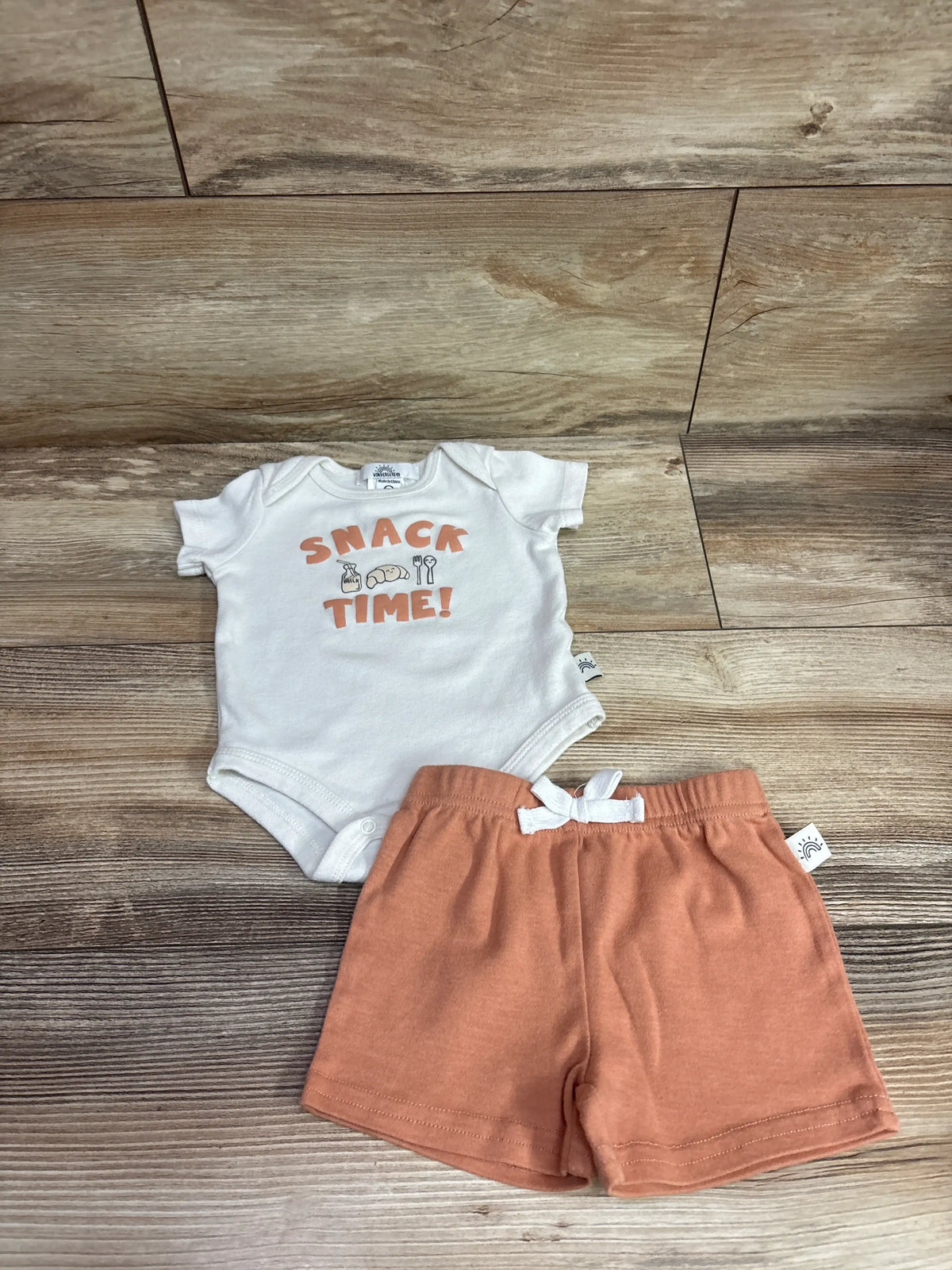 Top down view of Wonderluxe Snack Time bodysuit and shorts set showing white bodysuit and rust shorts on wood flooring