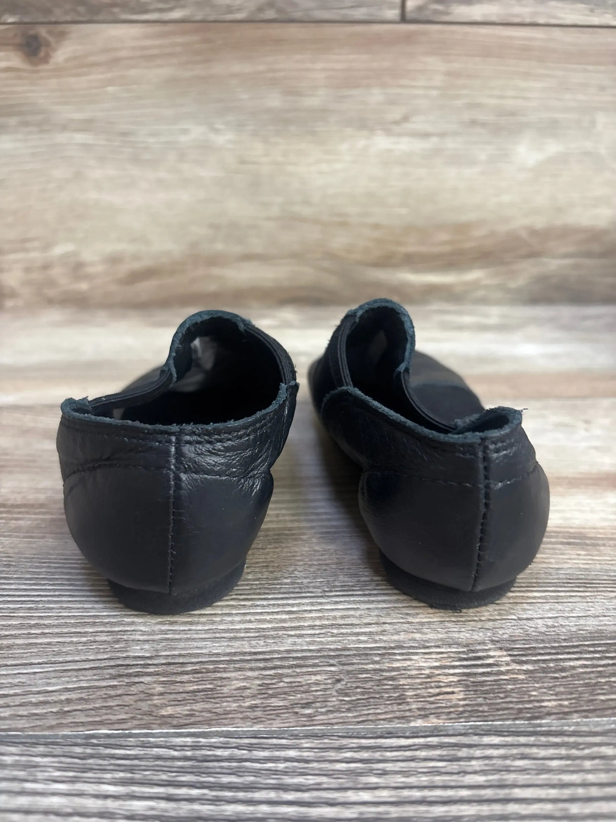 Rear view of Capezio E Series EJ2C girls’ black jazz slip-on shoes showcasing stitched heels and split-sole, on a wood floor background
