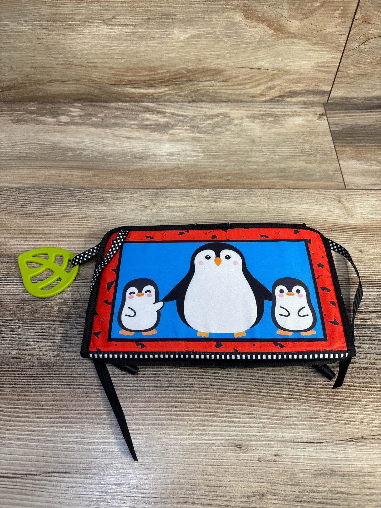 Flat lay of tummy-time cloth book showing high-contrast penguin cover, attached green teether, and polka-dot strap on wood floor