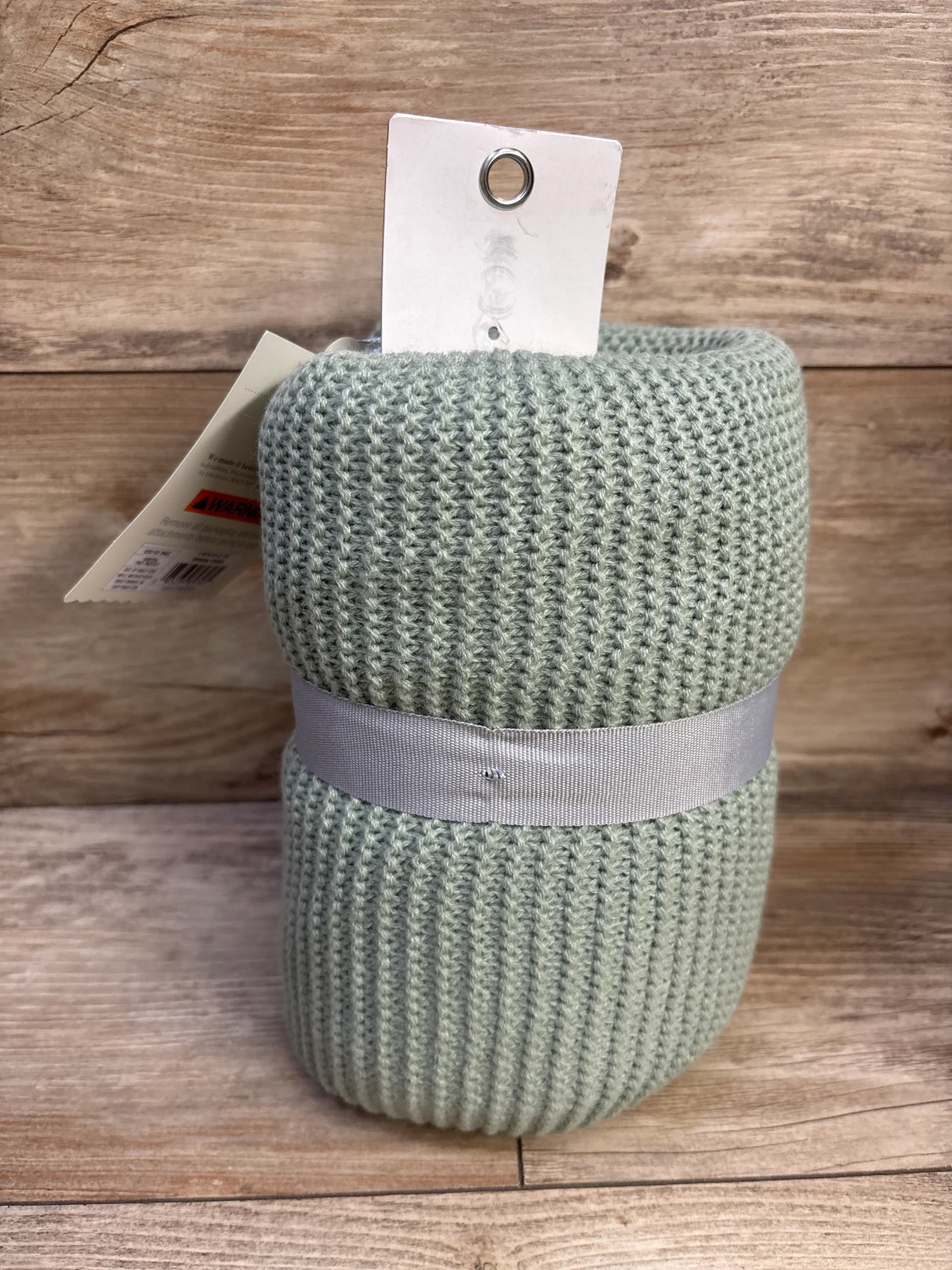 Close-up of rolled Cloud Island knit baby blanket in sage green showcasing ribbed knit texture and gray ribbon on wooden background