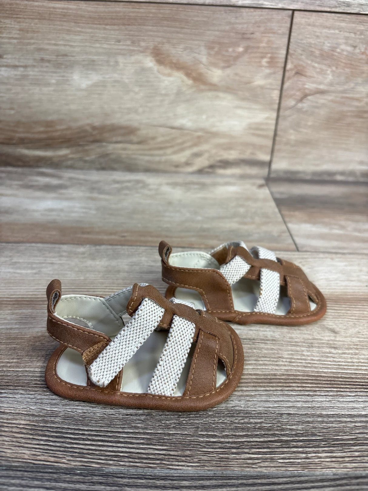 Angled view of Cat and Jack tan baby sandals showing white woven straps and brown faux-leather trim on woodgrain background