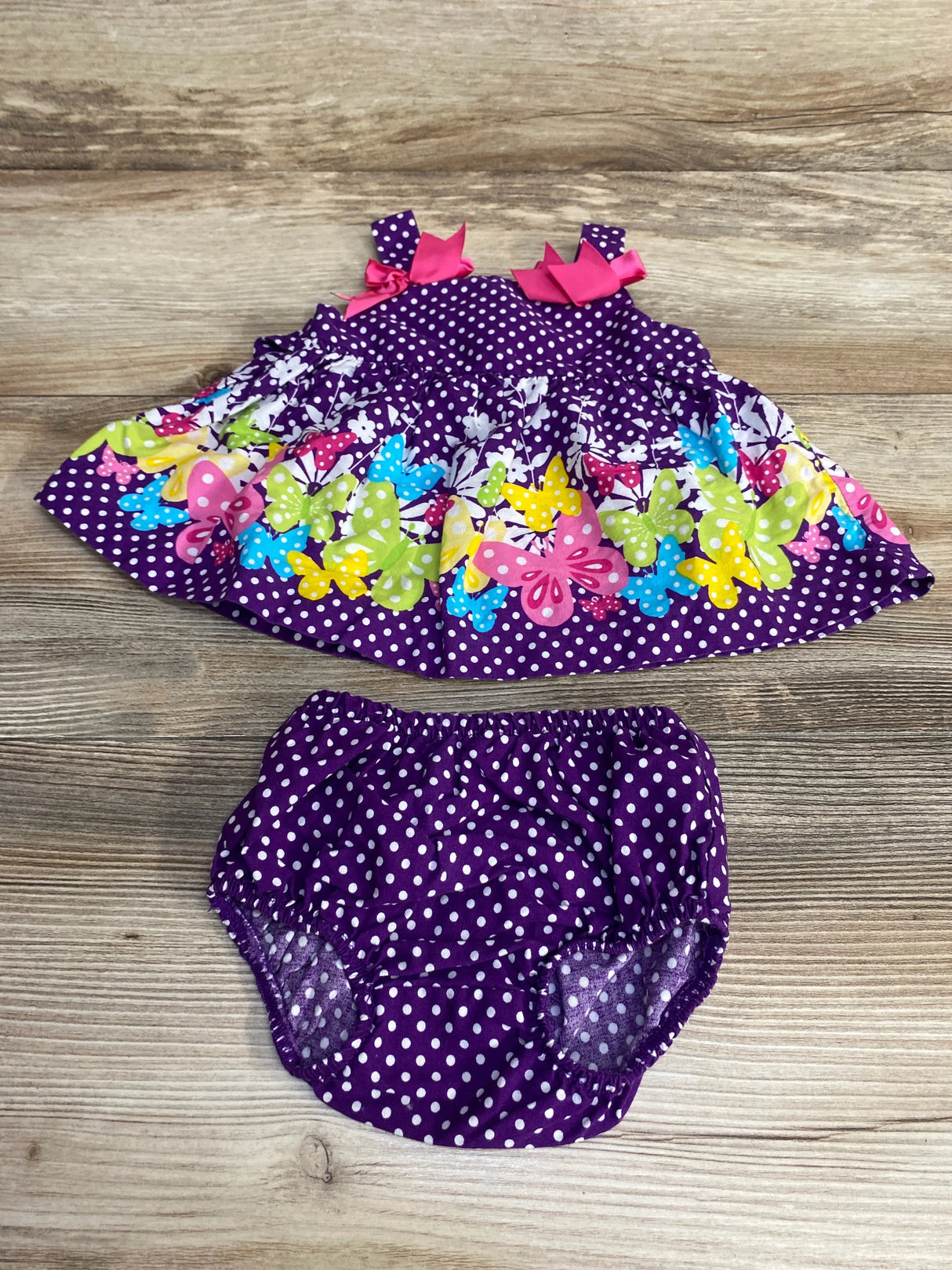 Flat lay of newborn purple polka-dot dress and matching bloomer showing multicolor butterfly border and pink bows on wood background