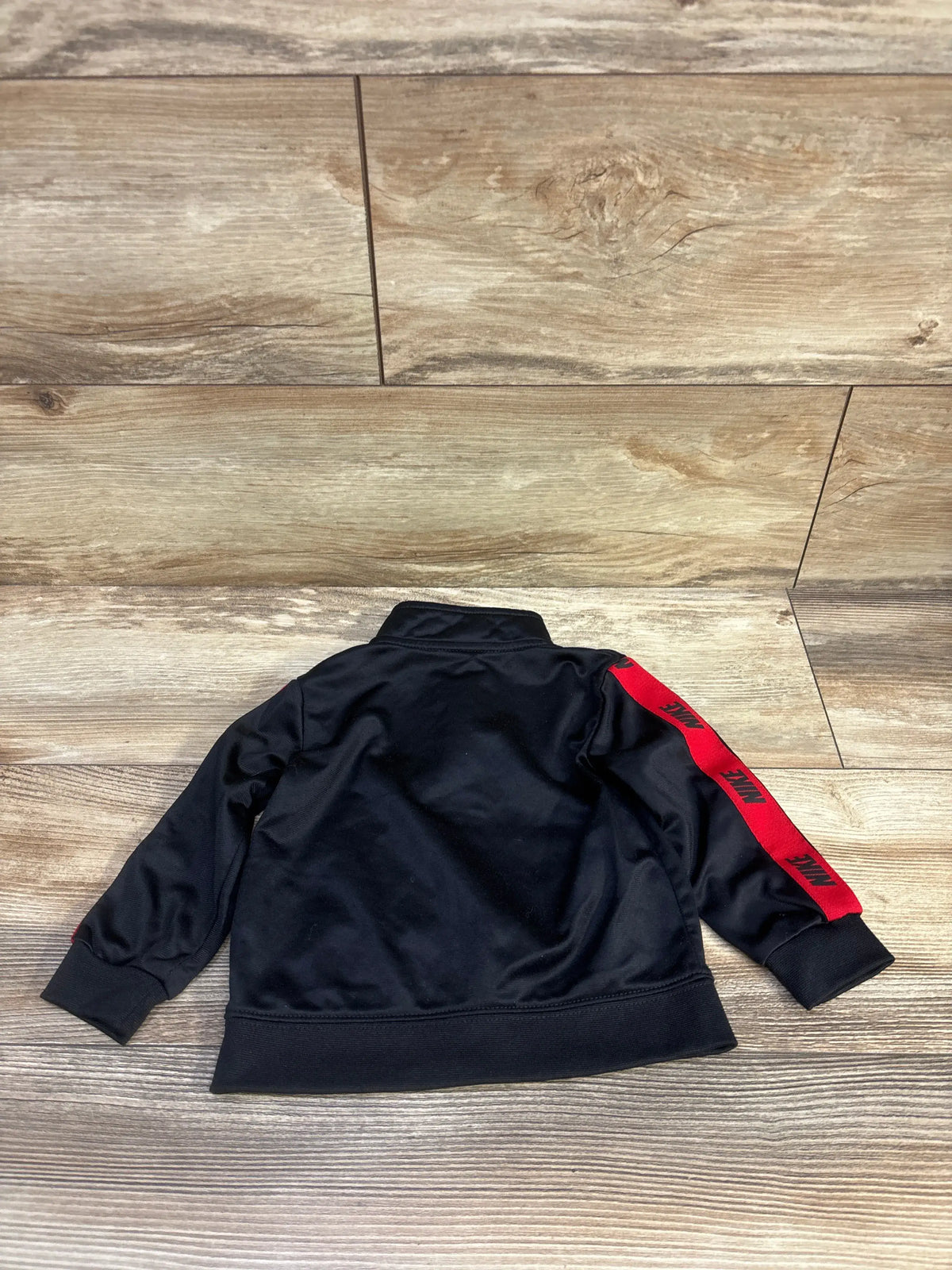 Back view of Nike toddler full-zip black track jacket showcasing red tape with repeated NIKE logos on sleeve on wood floor