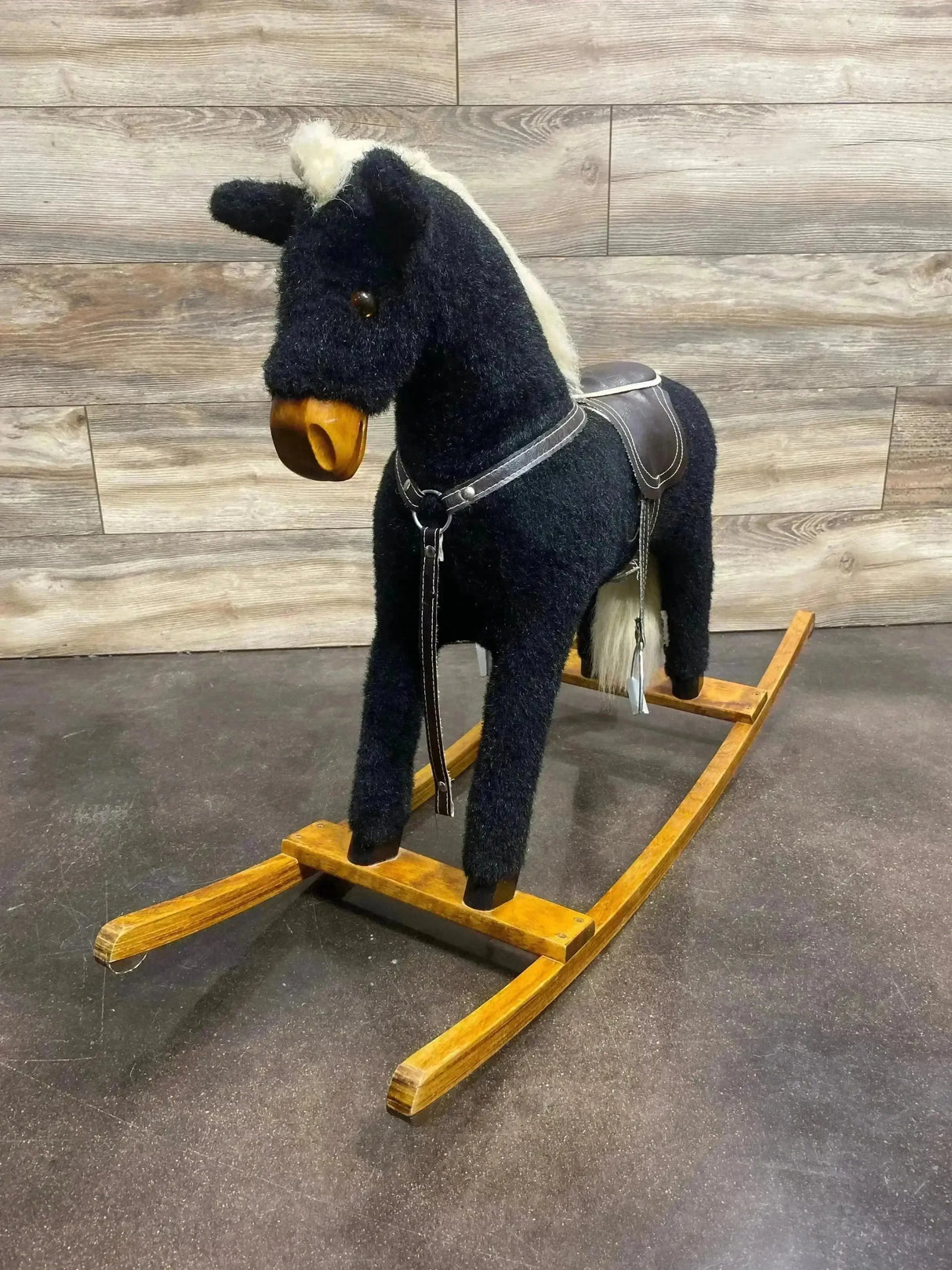 Three-quarter view of vintage black plush rocking horse with white mane, leather saddle and wooden curved rockers on gray floor