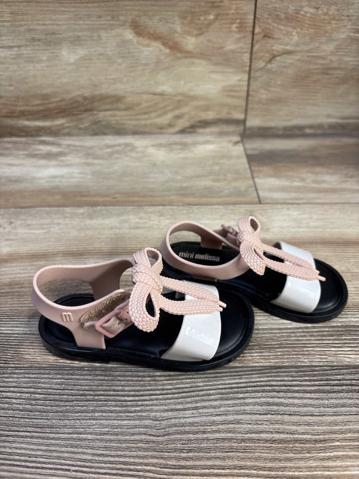 Angled view of Mini Melissa Mar toddler sandals showing pink braided bow, white patent toe strap, and black footbed on wood floor