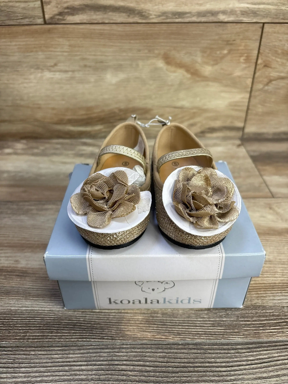 Front view of Koala Kids Sparkle Mary Jane flats with gold glitter, elastic strap and fabric rose on a Koalakids box