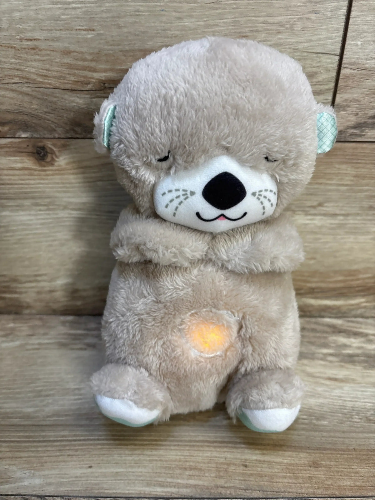 Front view of Fisher-Price Soothe ‘n Snuggle otter plush with embroidered face, crossed arms and glowing heart on wooden background