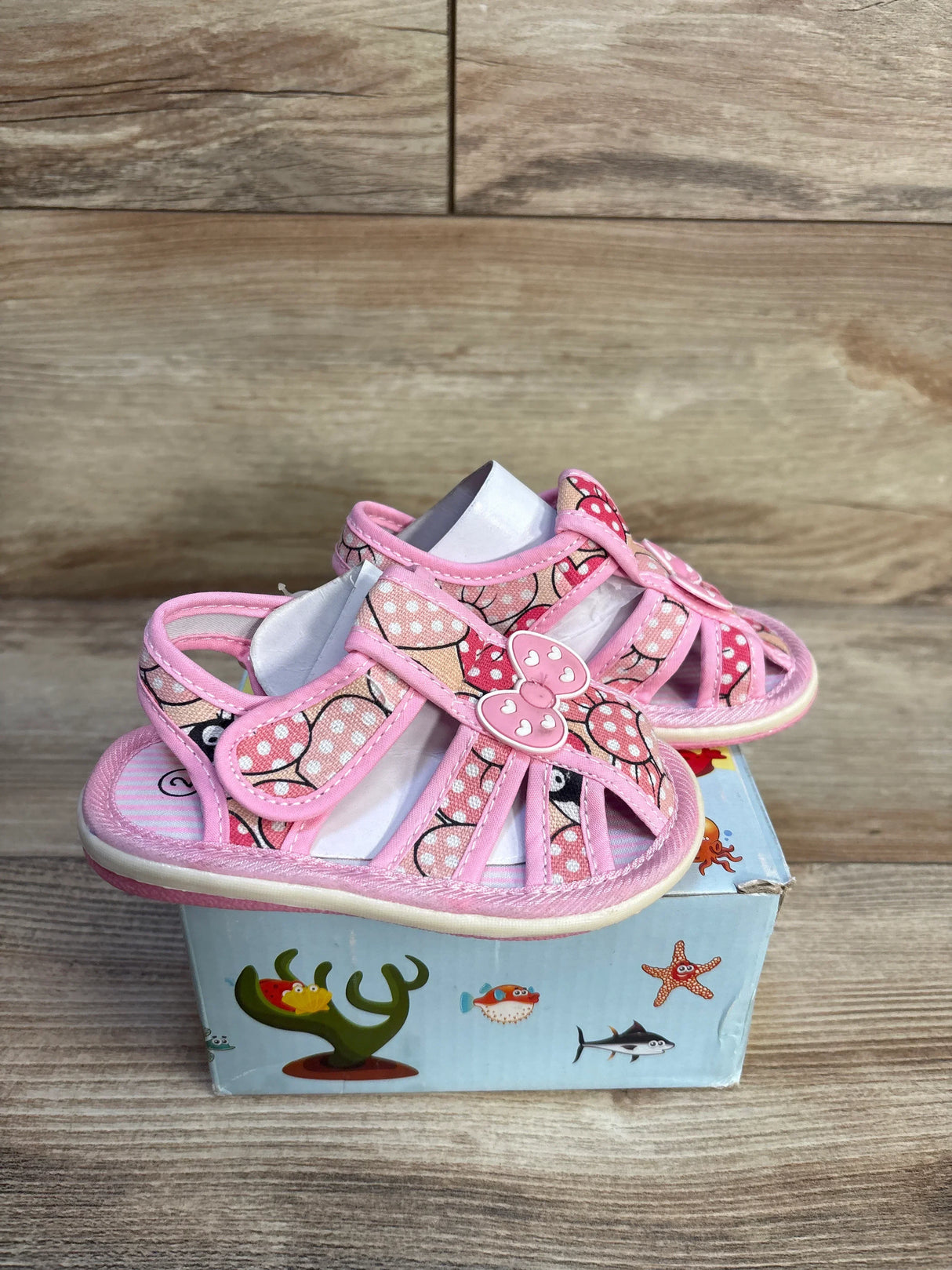 Pink toddler sandals with polka dots and bow accents, displayed on a decorated box