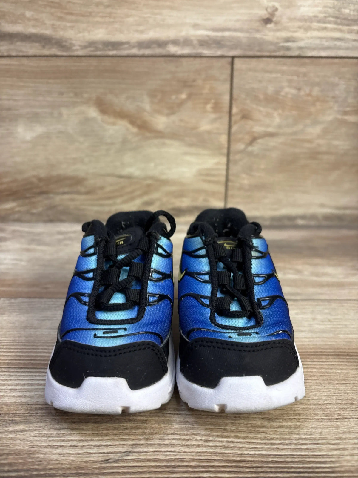 Front view of Nike Air Max Plus toddler sneakers showing blue gradient uppers, black trim, white soles, size 4C on wood floor background
