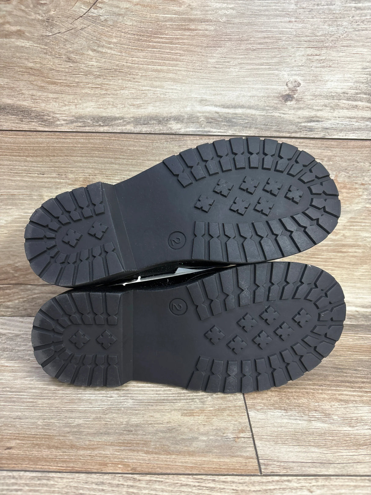 Pair of black rubber lug soles for kids' boots on wood floor, tread pattern visible