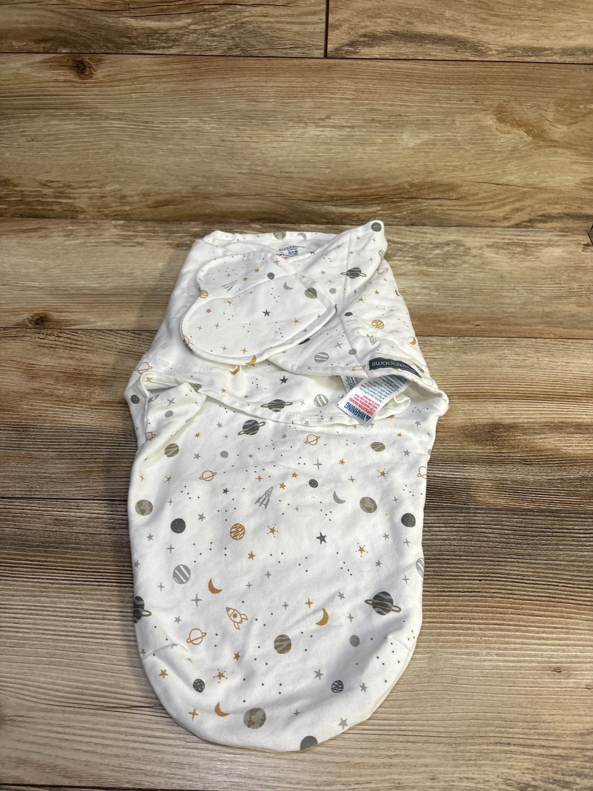 Flat lay of Swaddle Me swaddle wrap in white showing gray and gold planets and stars print on a wood background