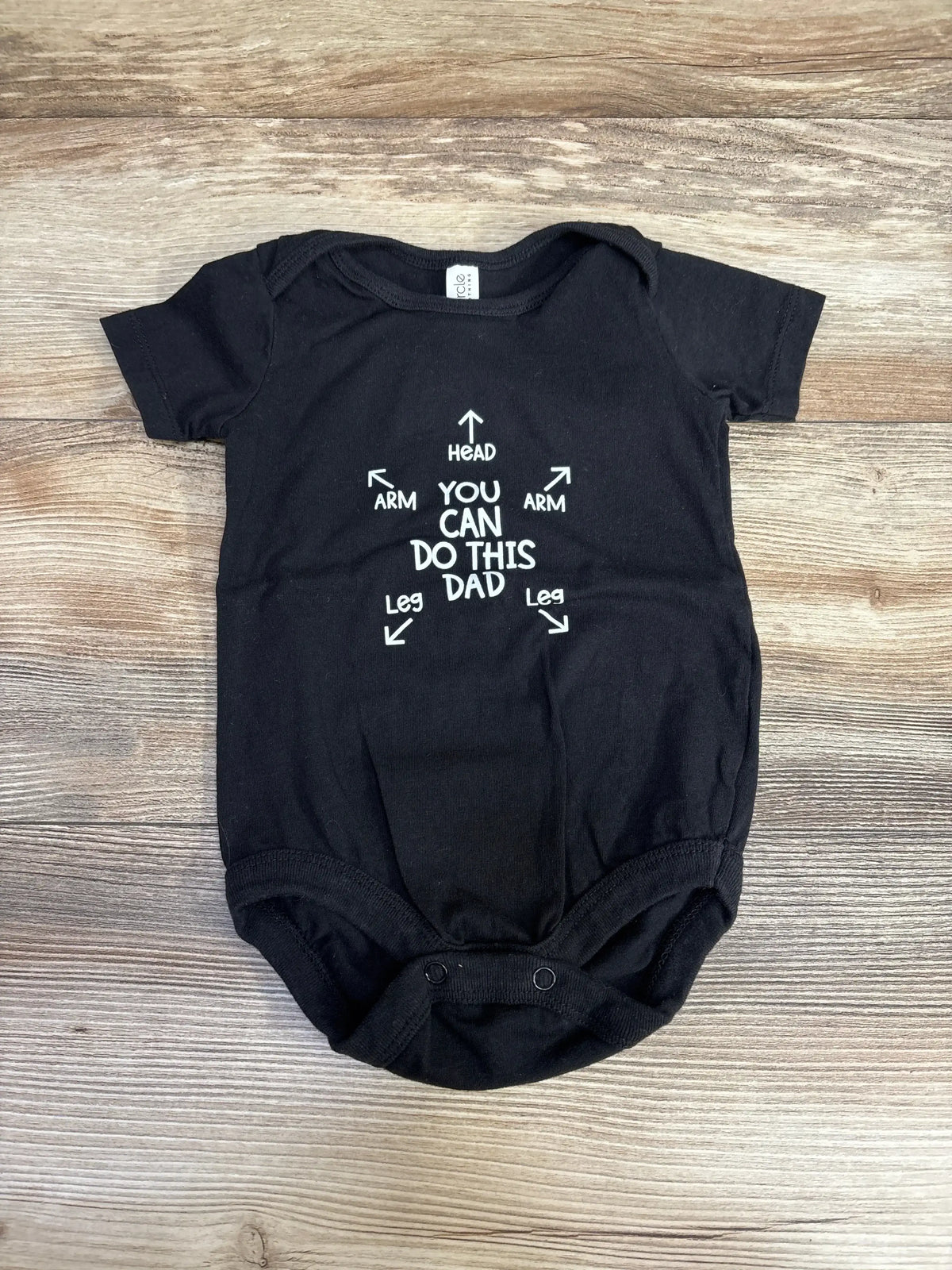 Front flat-lay of black 3-6M baby bodysuit showing white YOU CAN DO THIS DAD graphic with arrows on light wood background