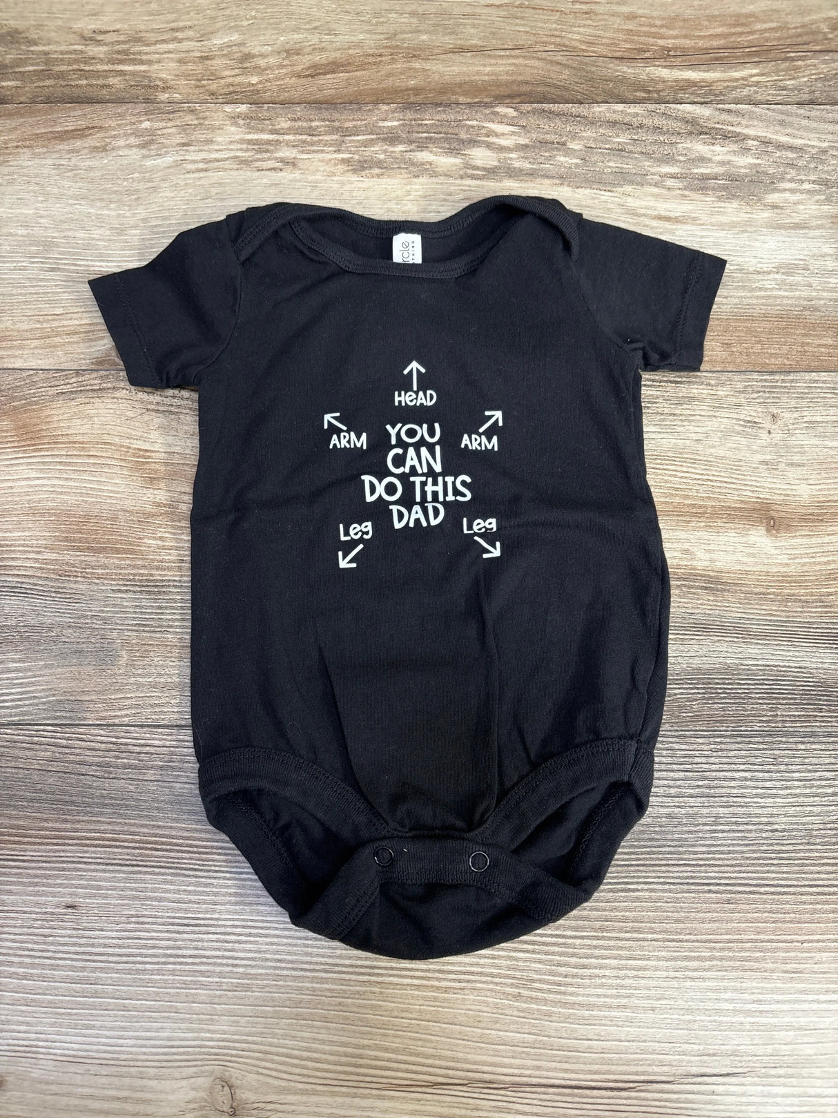Front flat-lay of black 3-6M baby bodysuit showing white YOU CAN DO THIS DAD graphic with arrows on light wood background