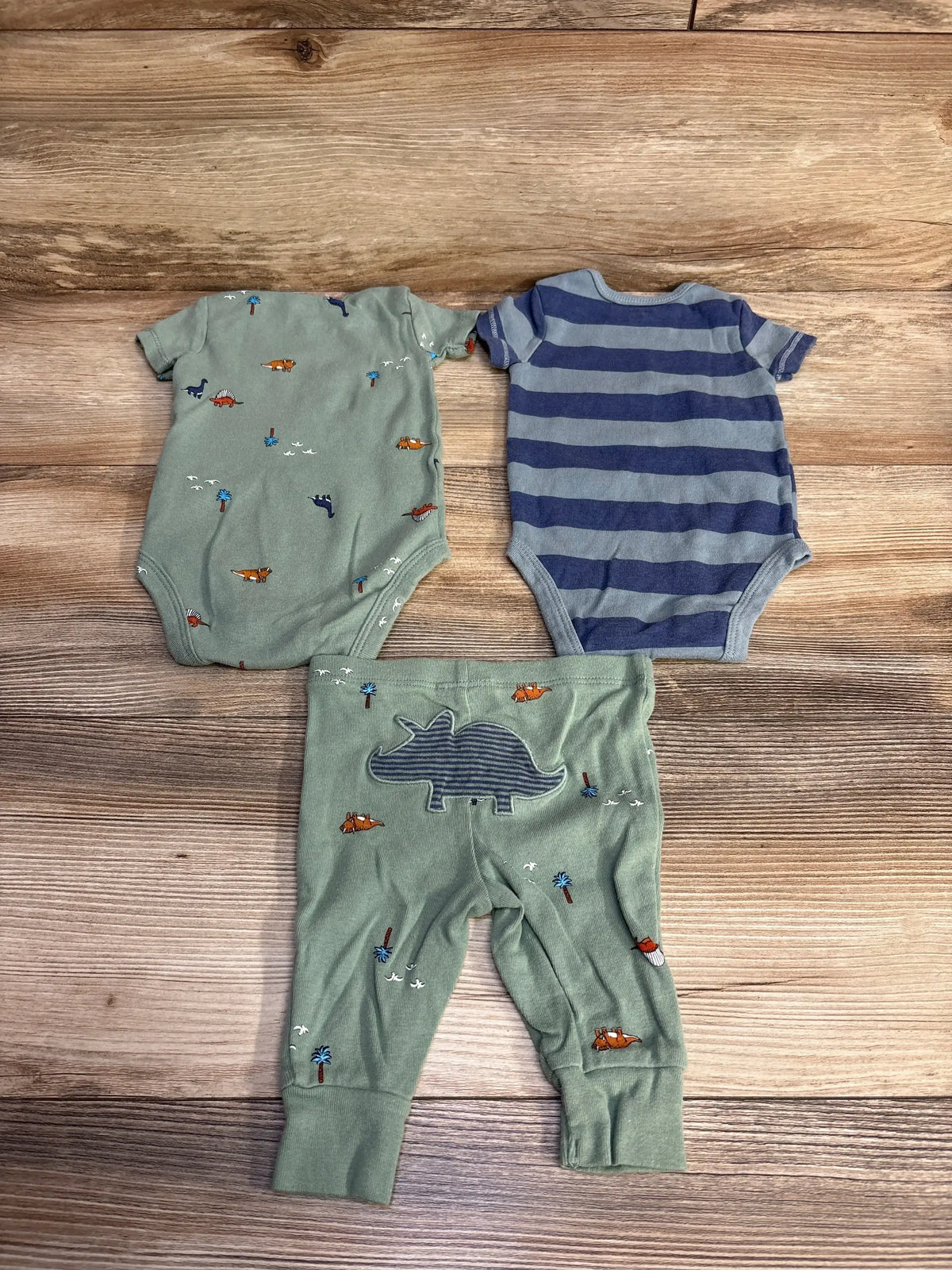 Flat lay of 3-piece dinosaur baby bodysuit set with green dinosaur print, blue stripes, and matching pants on wood background