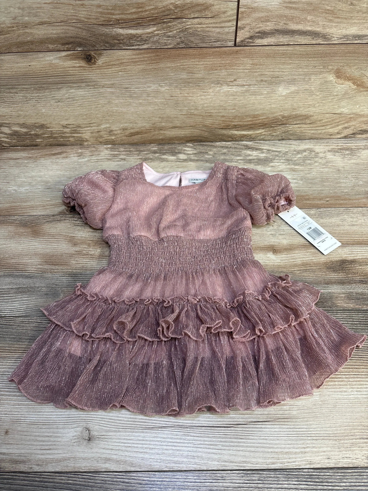 Flat lay of Habitual Kids Sparkle Holiday dress size 12 months showing pink shimmer fabric, puff sleeves, smocked waist, and tiered ruffle skirt on wooden floor