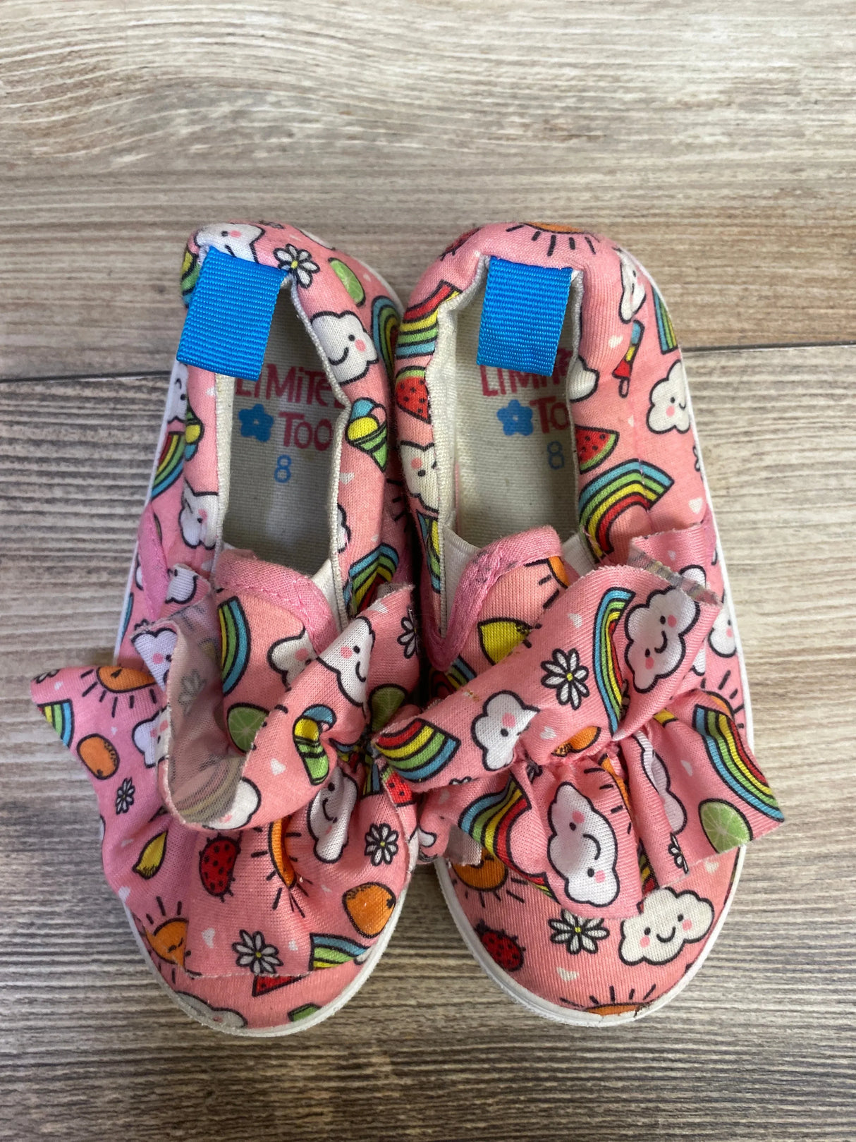Top down view of Limited Too pink slip on toddler sneakers with fabric bows and rainbow cloud print on wood floor size 8C