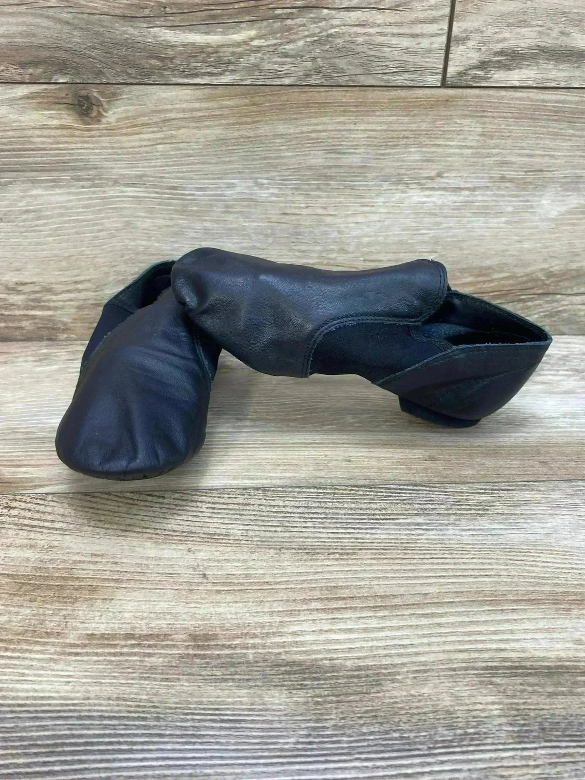 Side view of Capezio E-Series black jazz slip-on shoes showing leather upper, elastic sides, size 8C on light wood background