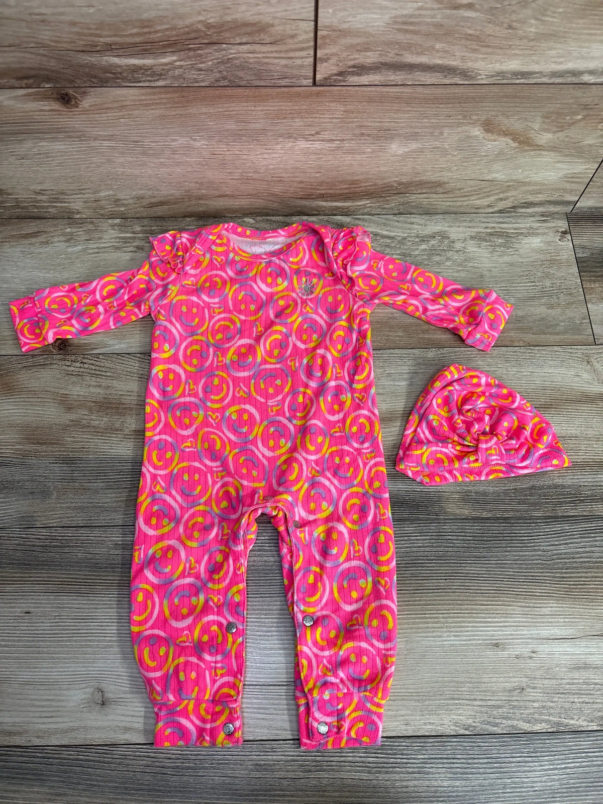 Pink baby romper and hat set with yellow smiley face print, laid on wood floor